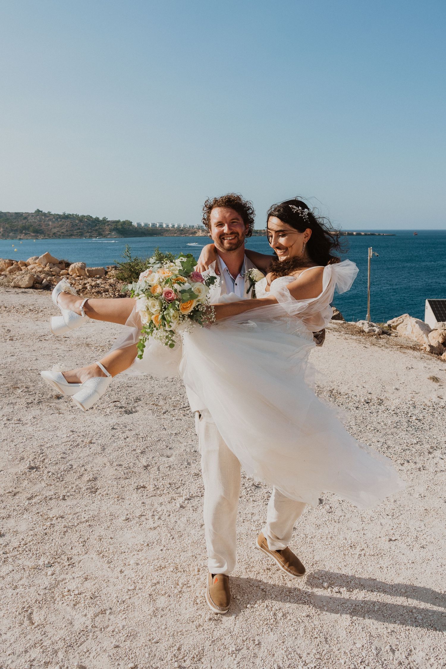 A joyful wedding couple shares a playful moment as they dance on a coastal cliff.