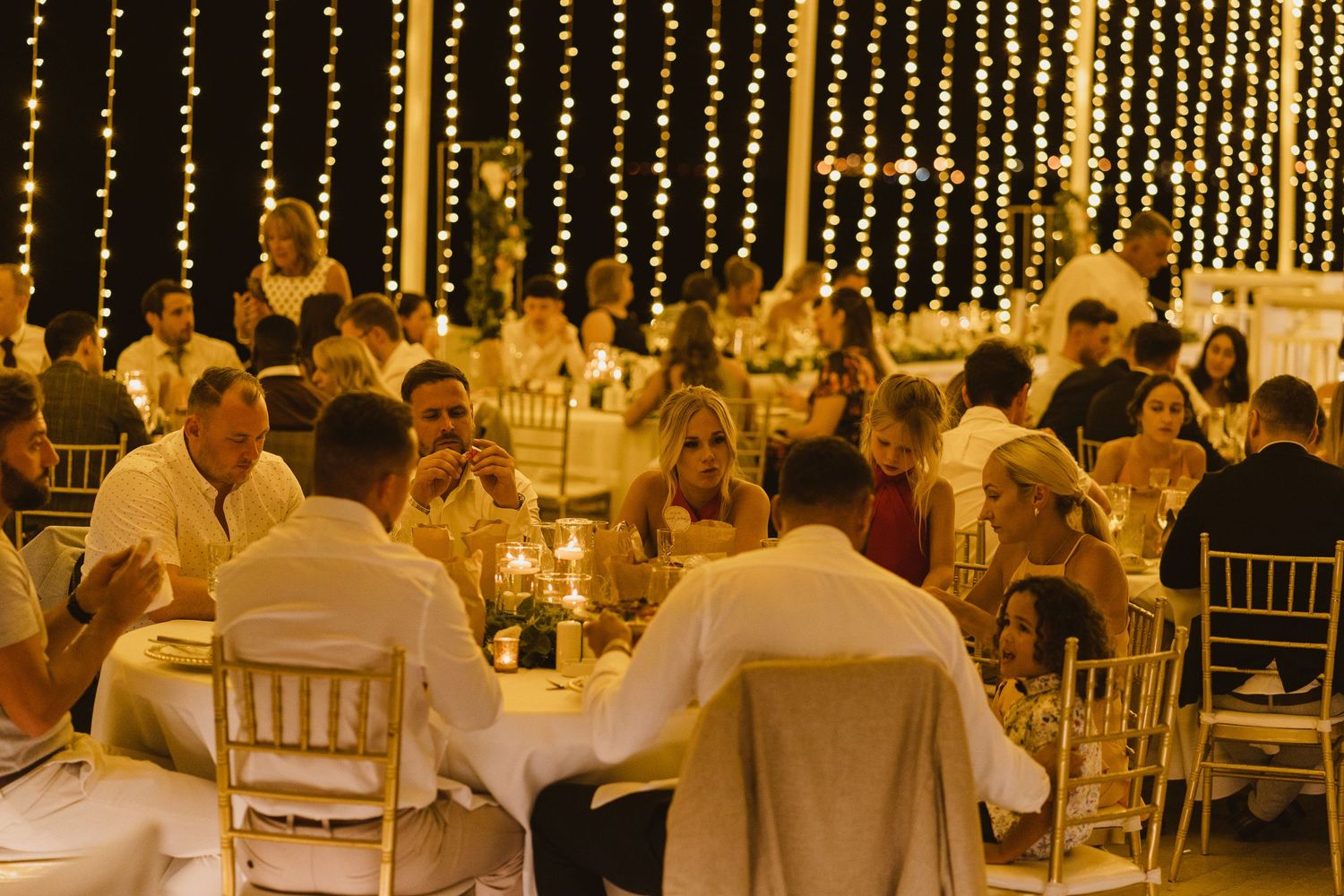 Wedding guests seated at round tables under warm glowing string light canopy.