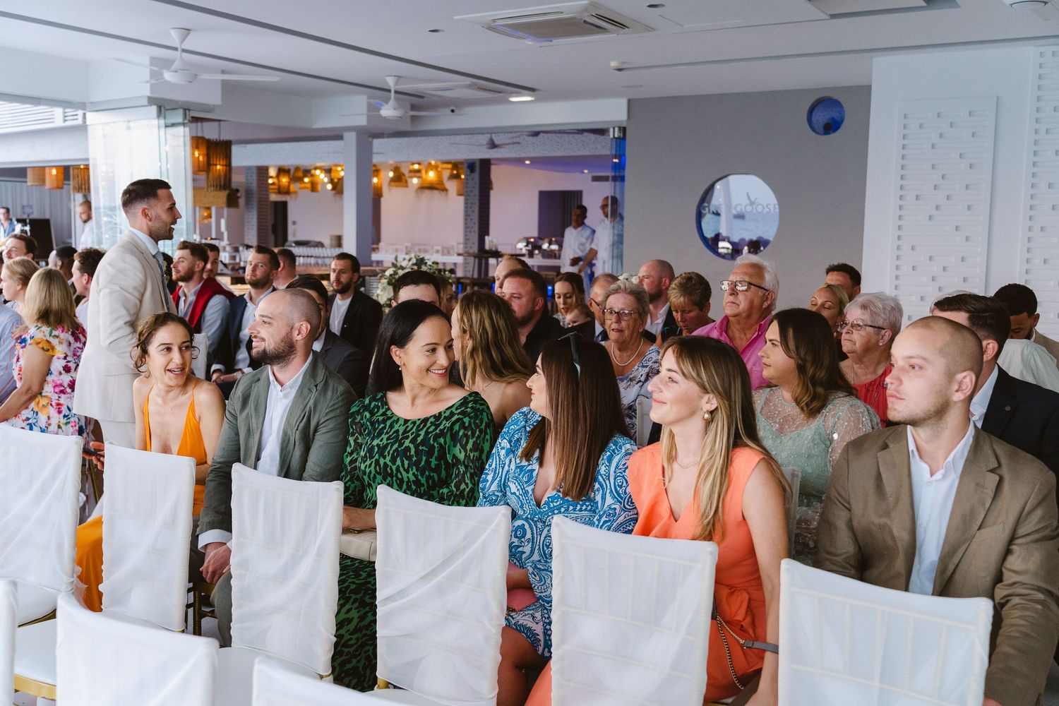 Wedding ceremony guests seated in white chairs at an indoor venue.