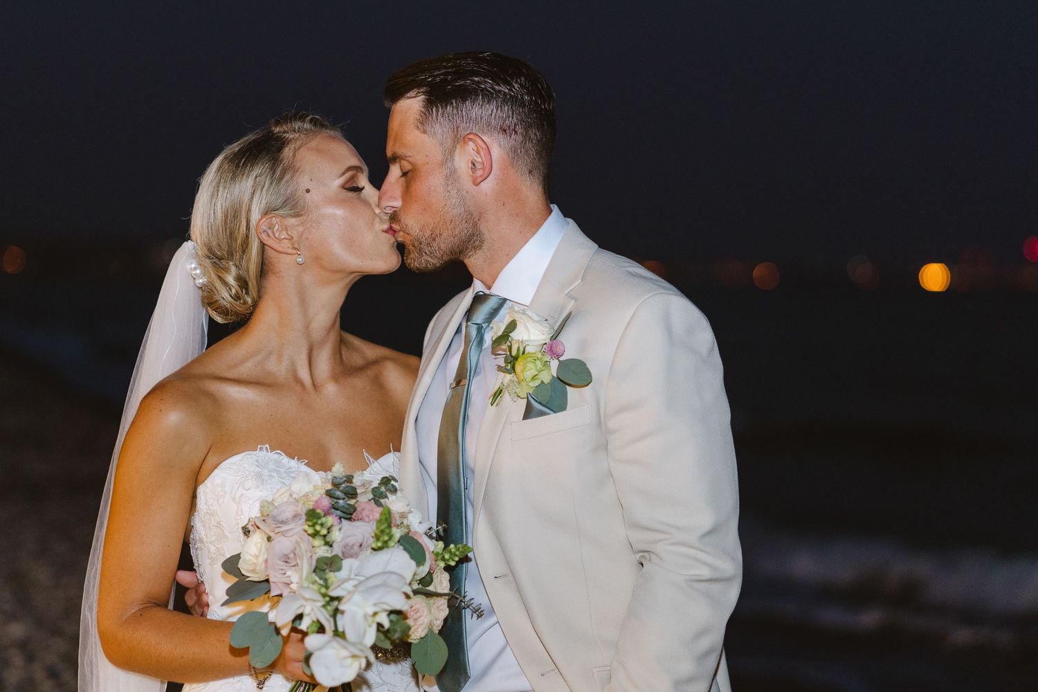 Newlyweds share a romantic kiss on a rocky beach at night with city lights in the background.