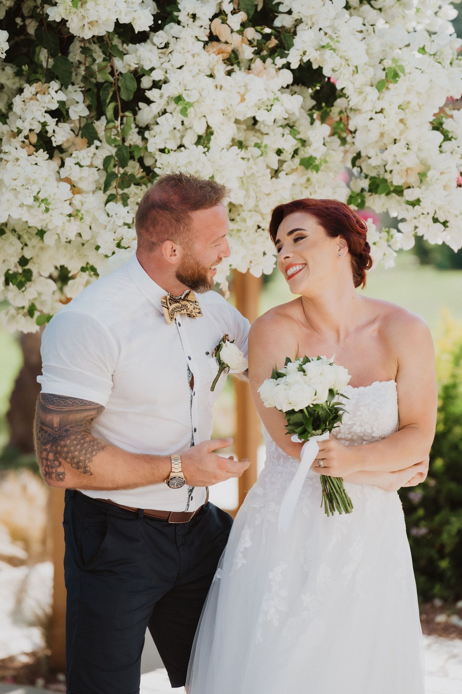 A happy couple shares laughter during their wedding photoshoot beneath white flowering trees.