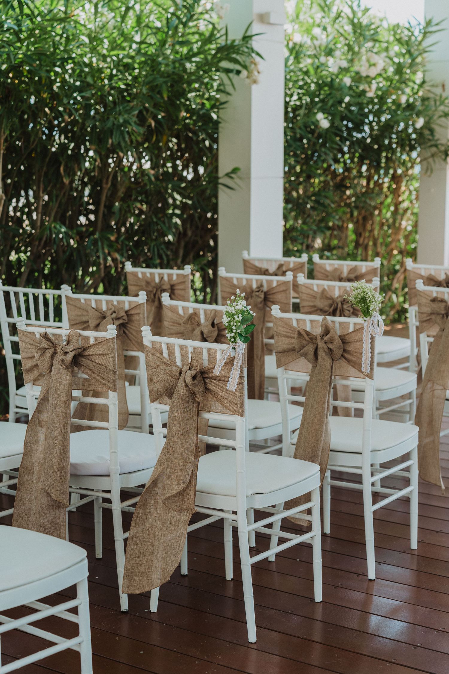 White wedding chairs with burlap sashes arranged on wooden deck for outdoor ceremony.