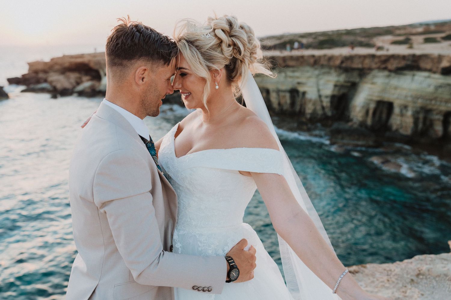 A romantic wedding couple embraces near stunning turquoise waters and rocky coastal cliffs at sunset.