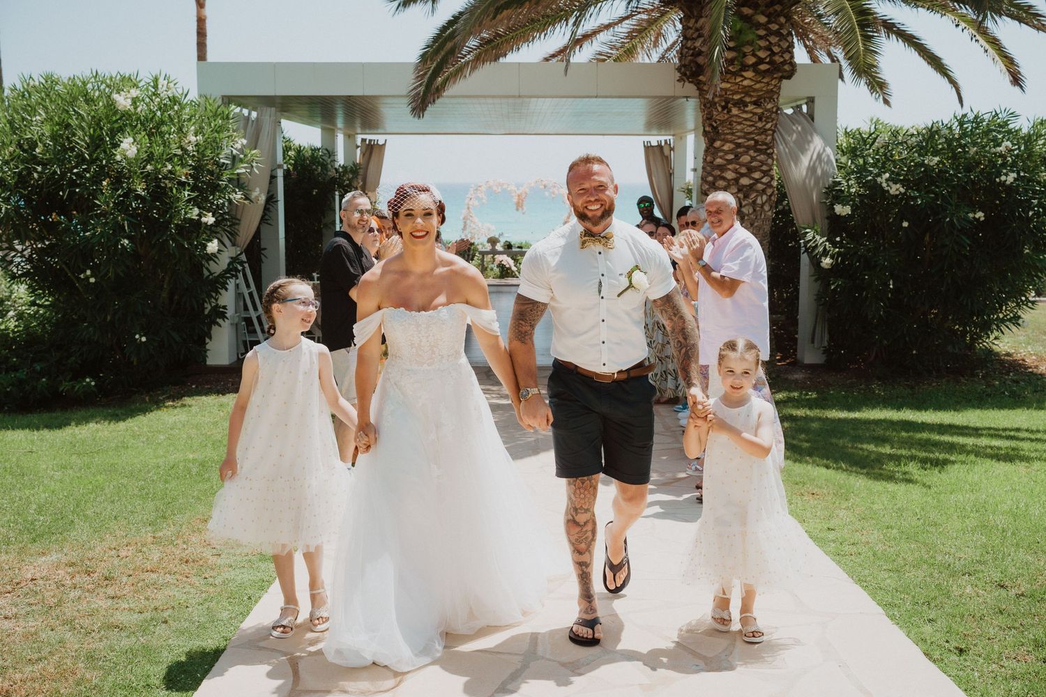 Wedding party walks along a sunny garden path with palm trees.