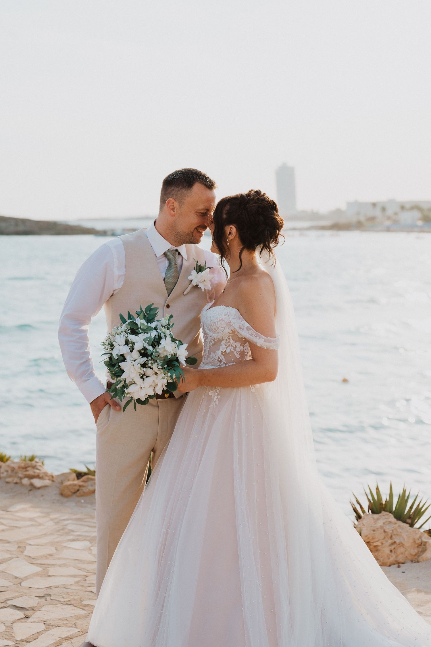 A romantic coastal wedding photo with a couple in beige and white attire holding a eucalyptus bouquet by the ocean.