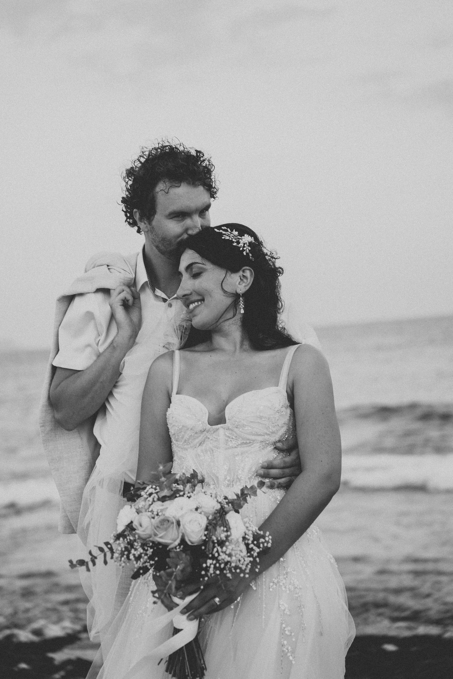 Black and white wedding portrait showing tender moment between couple by the ocean.