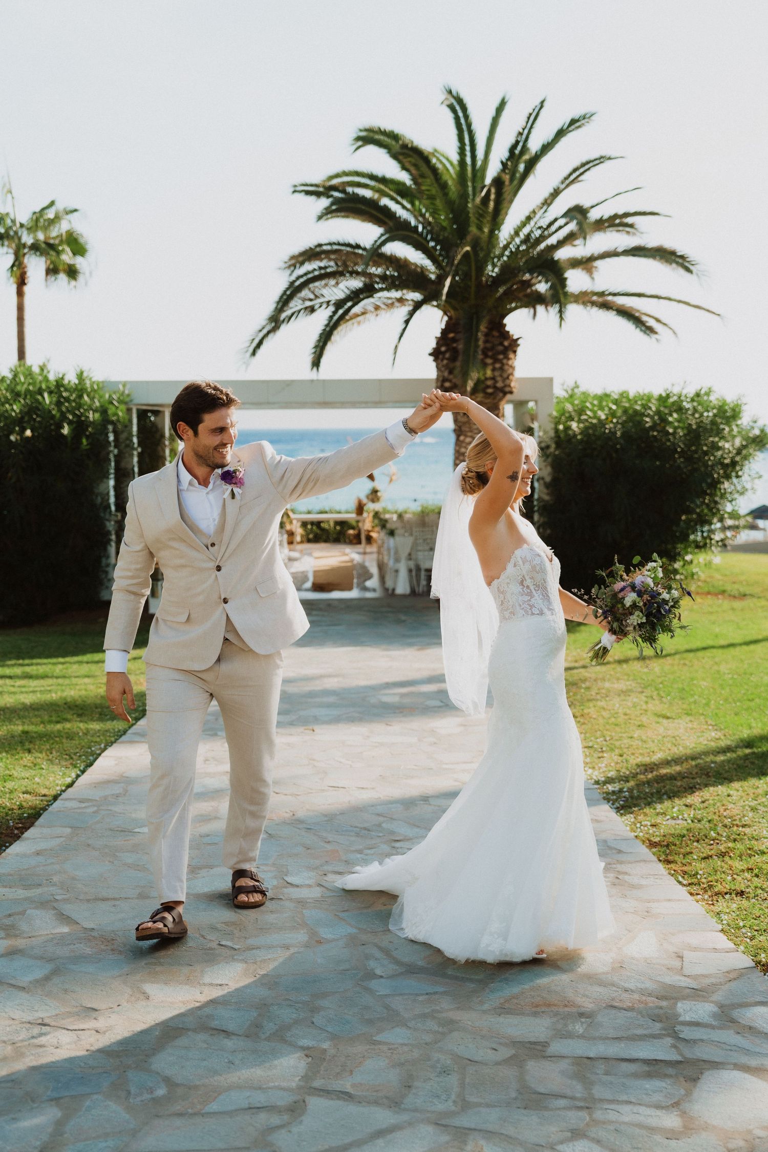 A couple in wedding attire dances playfully on a stone patio with palm trees in the background.