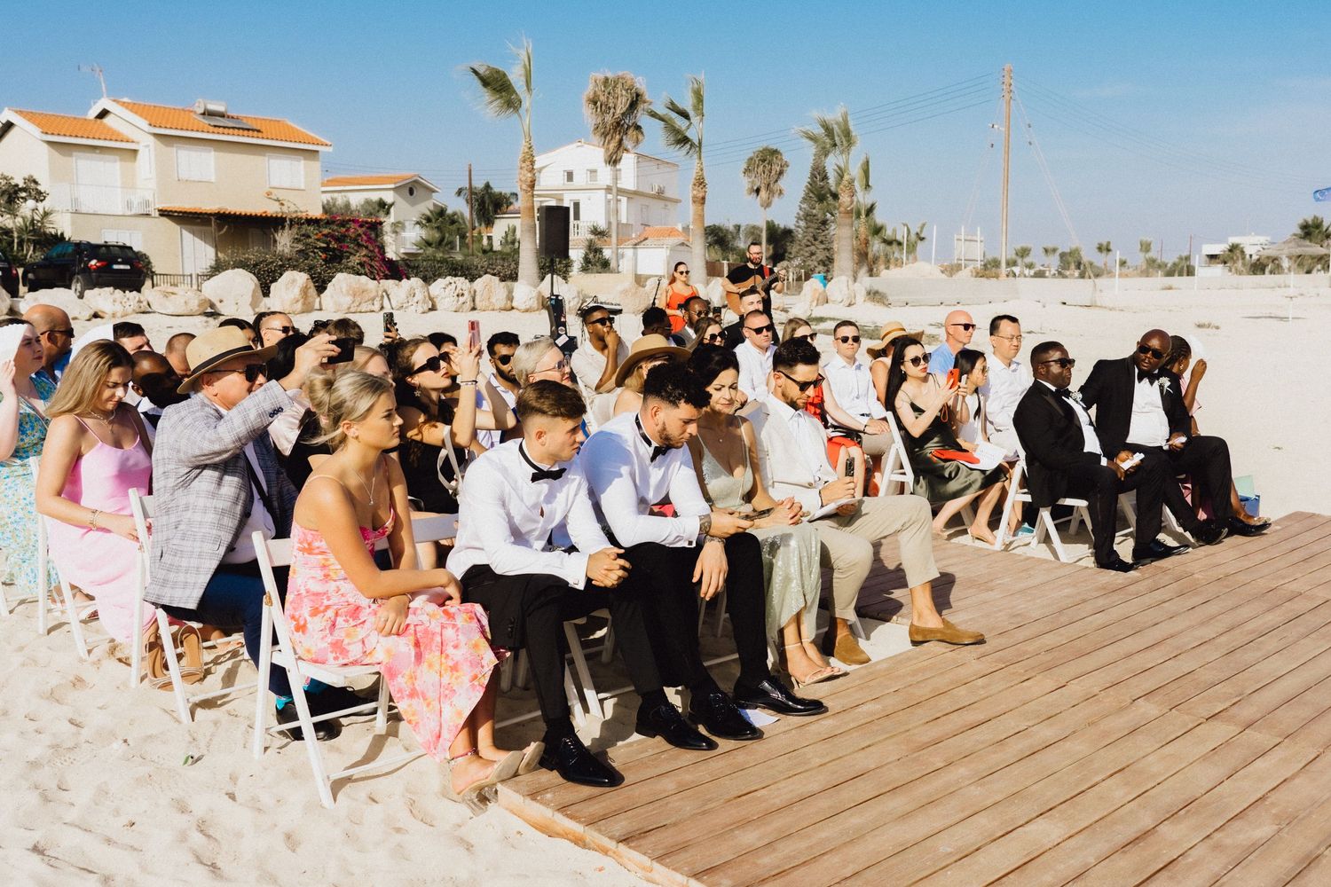 Wedding guests seated on wooden boardwalk at beachside ceremony.