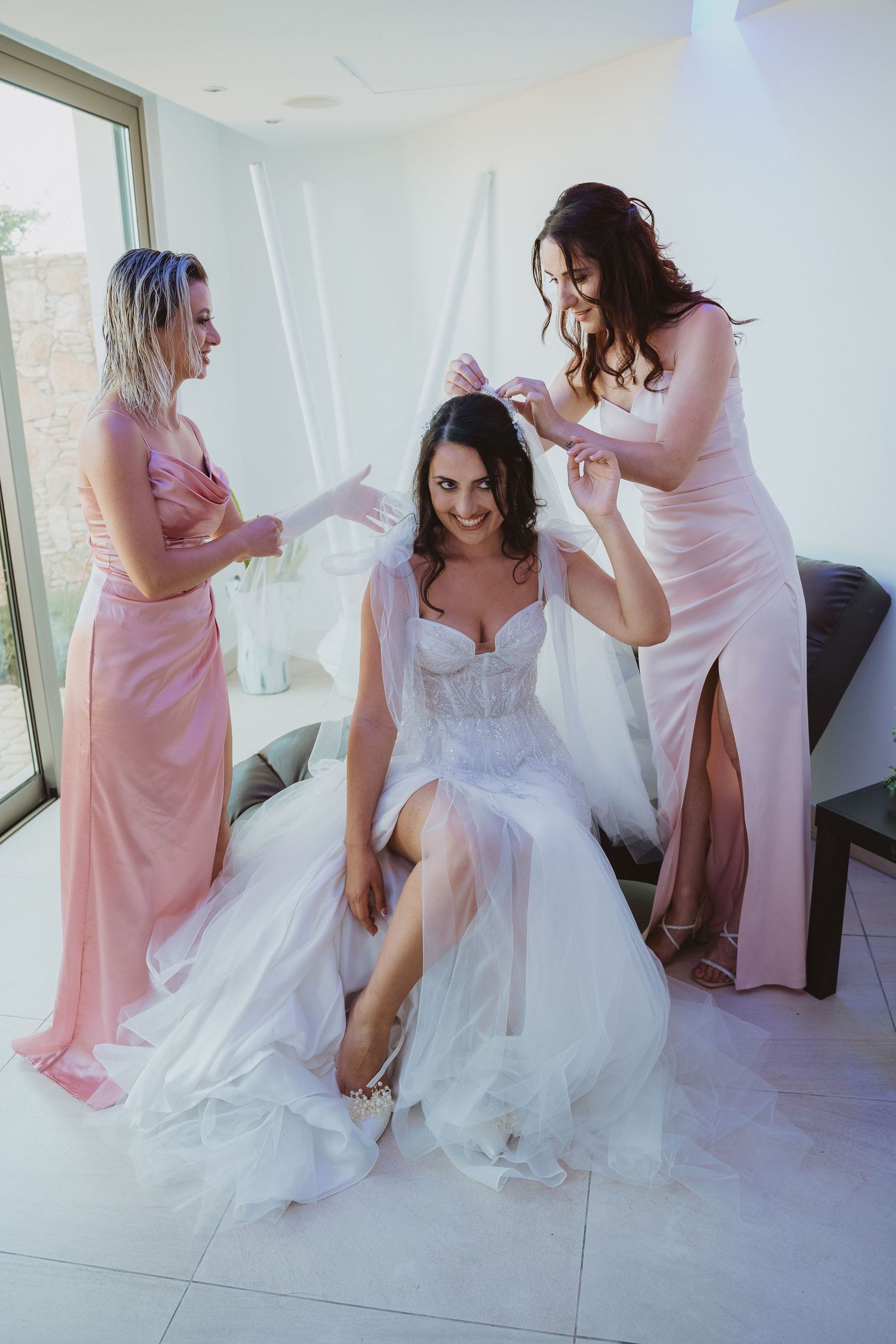 Bridesmaids in pink dresses help the bride with preparations in a bright modern room.