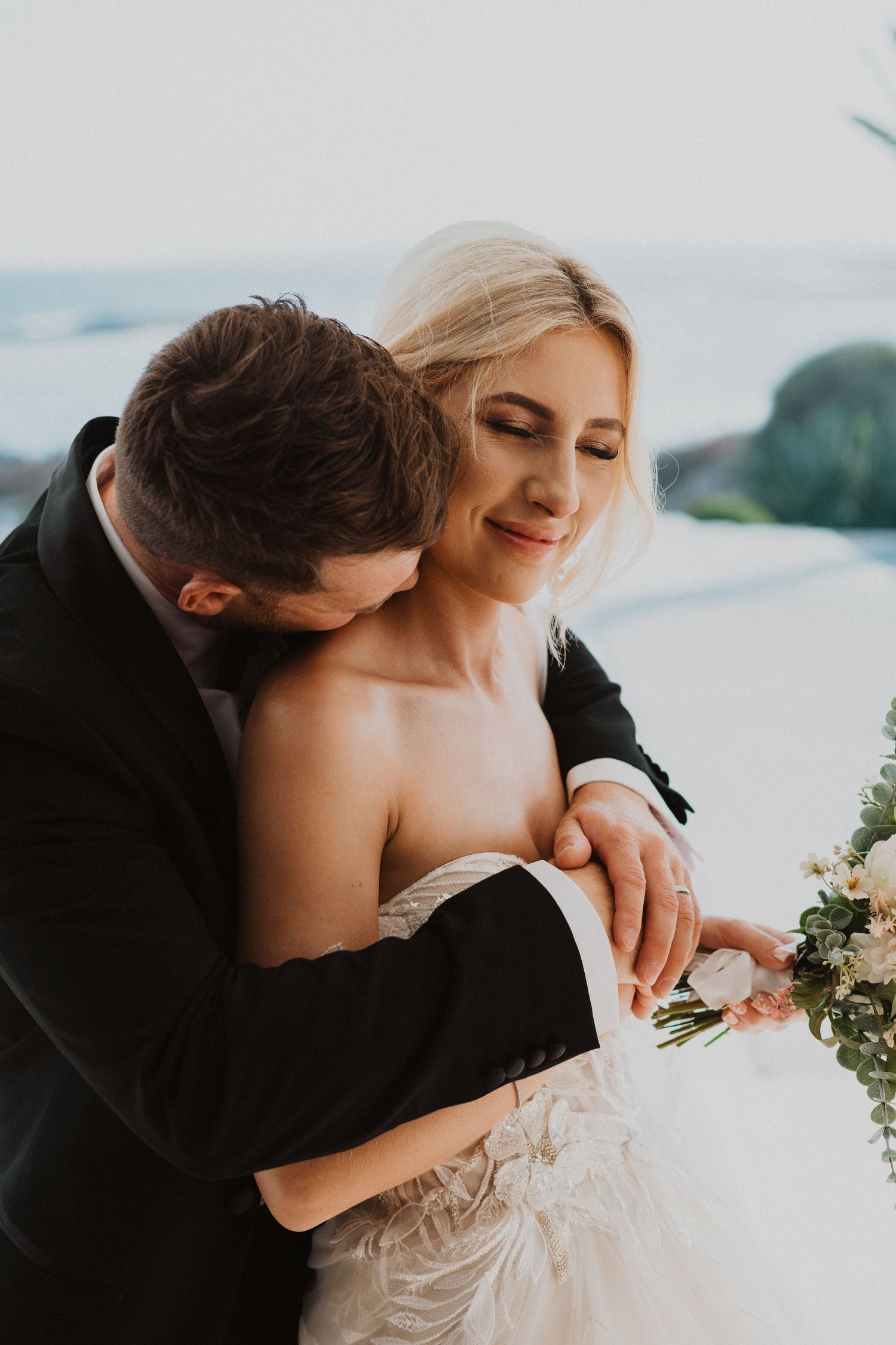 Tender embrace between newlyweds with coastal backdrop and white bouquet.