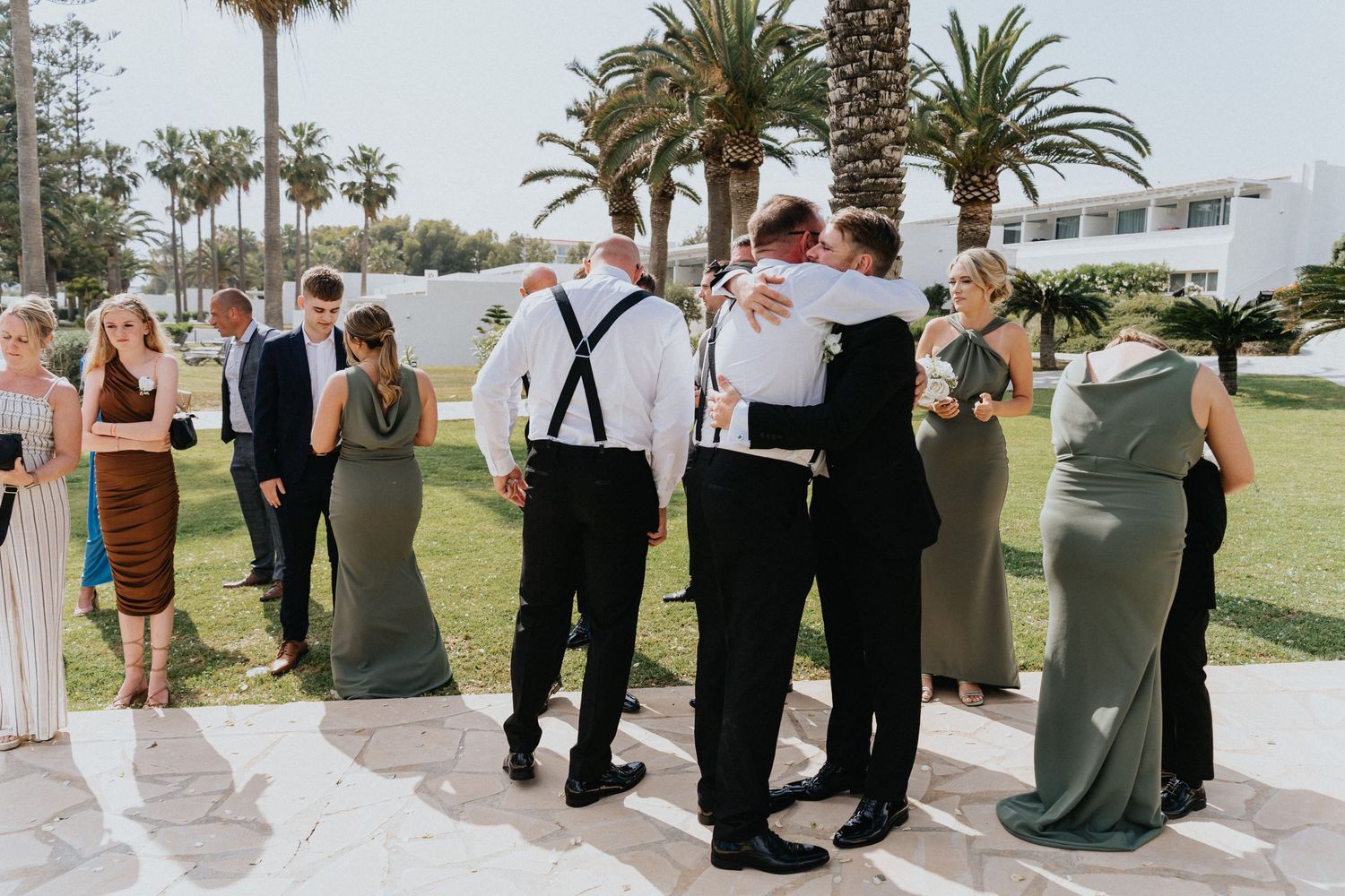 Groomsmen in suspenders and wedding guests mingle during outdoor reception by palm trees.