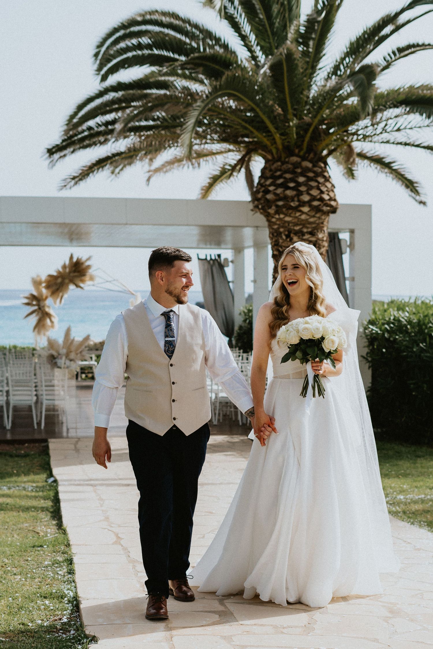 A wedding couple walking hand in hand on a pathway with a palm tree and outdoor venue in the background.