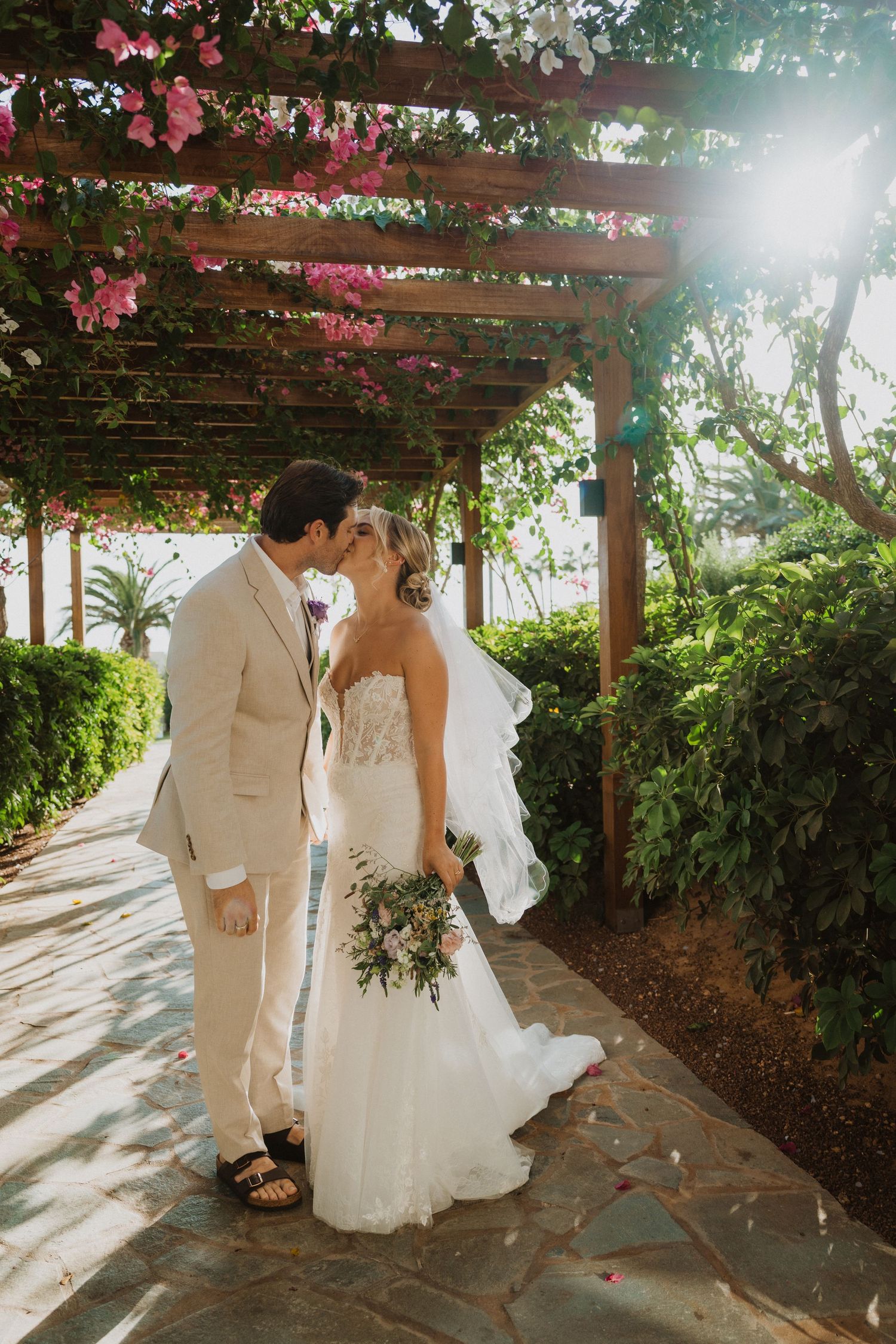 Sunlit wedding portraits under wooden pergola covered in pink flowers and vines.