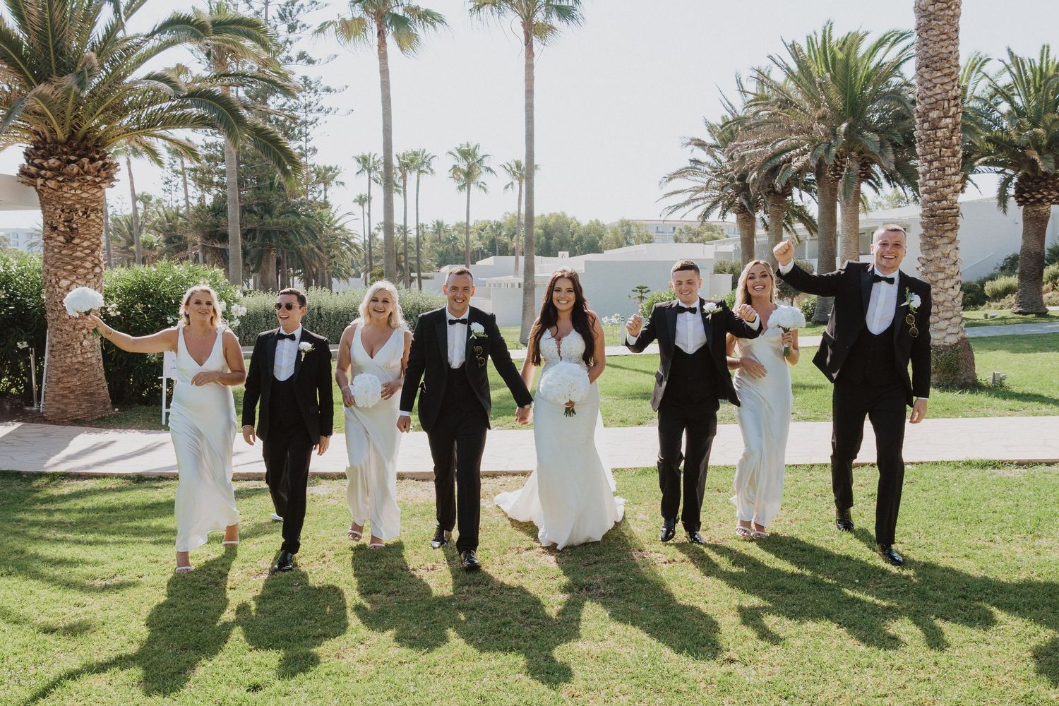 Wedding party walking and posing playfully along a palm tree-lined path outdoors.