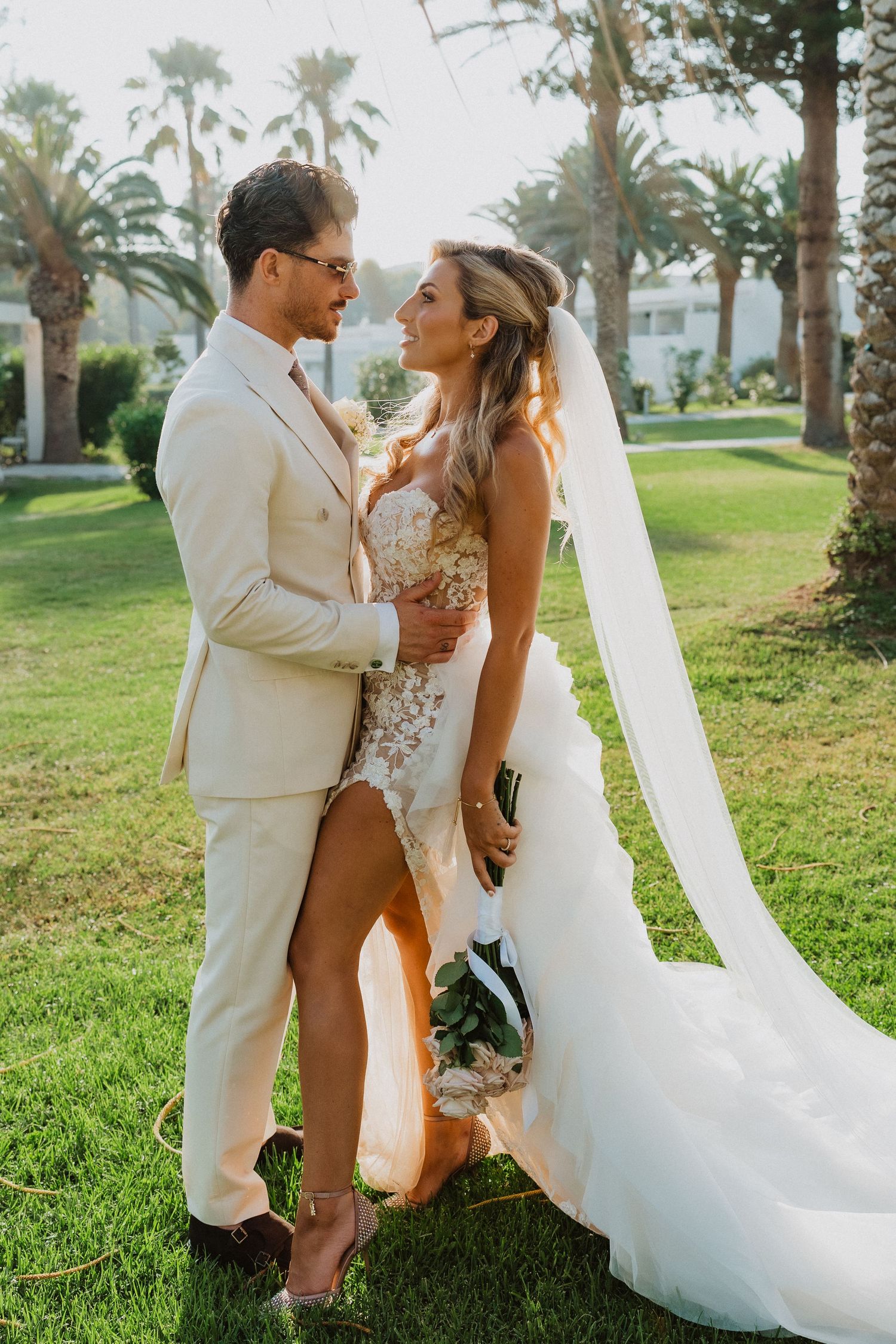 A romantic wedding couple embraces outdoors on a grassy lawn with palm trees in the background.
