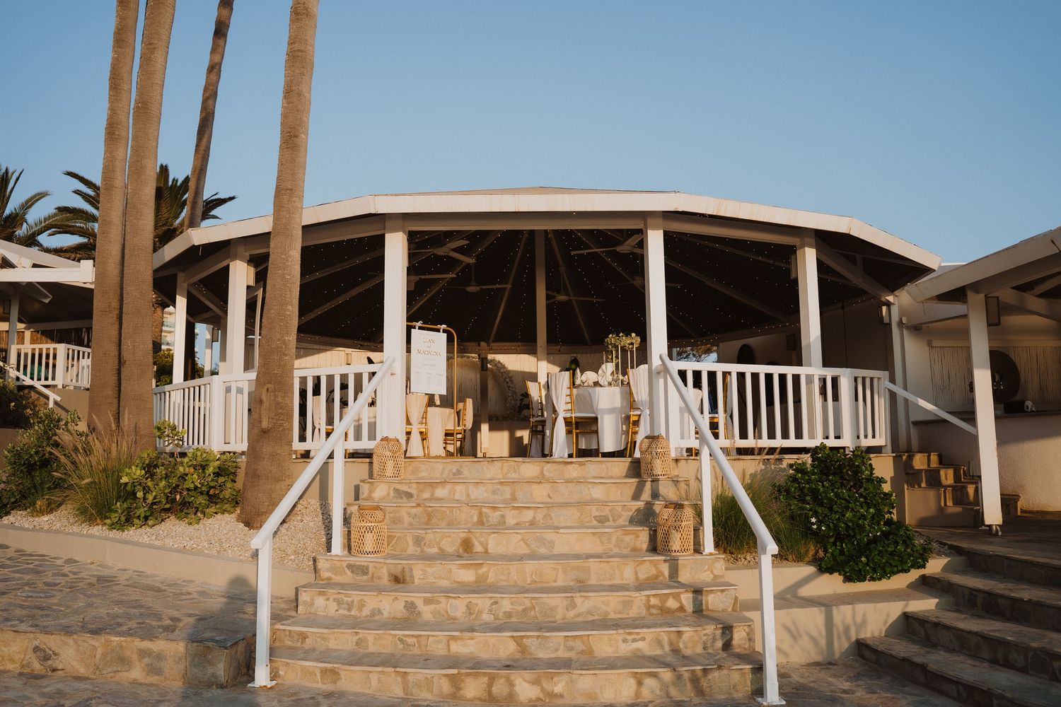 Elegant beachside wedding venue with white columns, wraparound deck, and stone staircase at sunset.