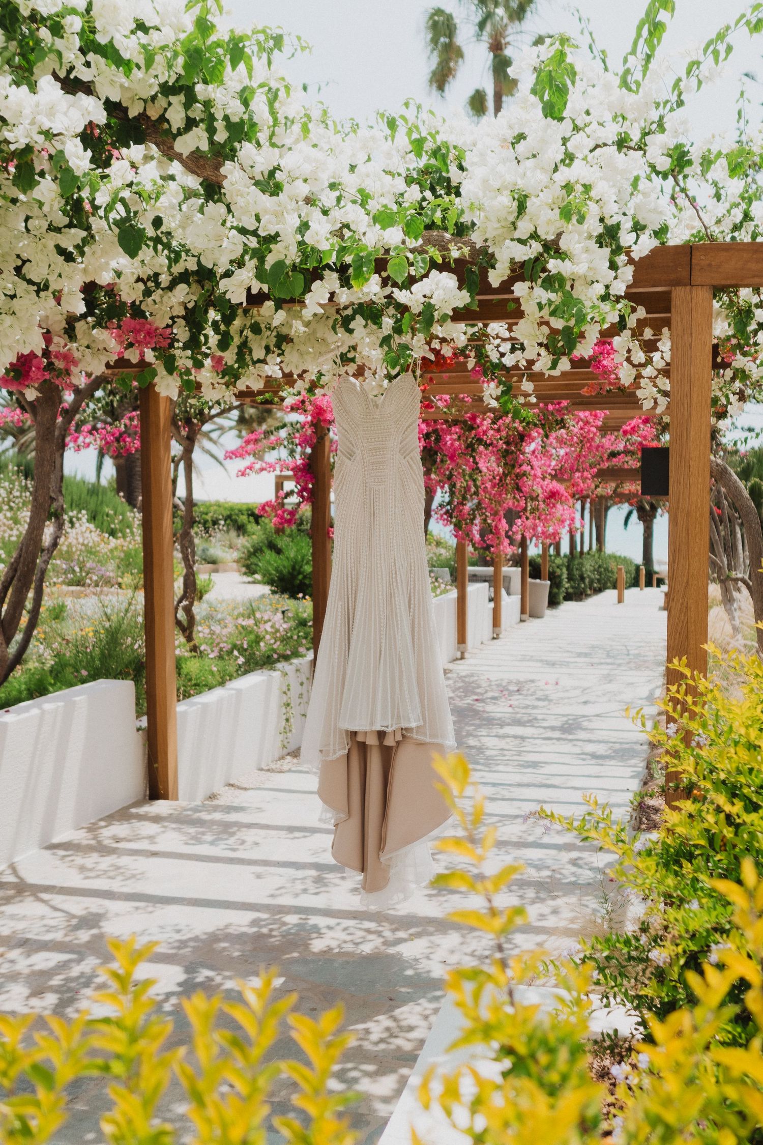 A elegant wedding dress hanging under a floral pergola with white and pink bougainvillea blooms lining a sunny garden pathway.