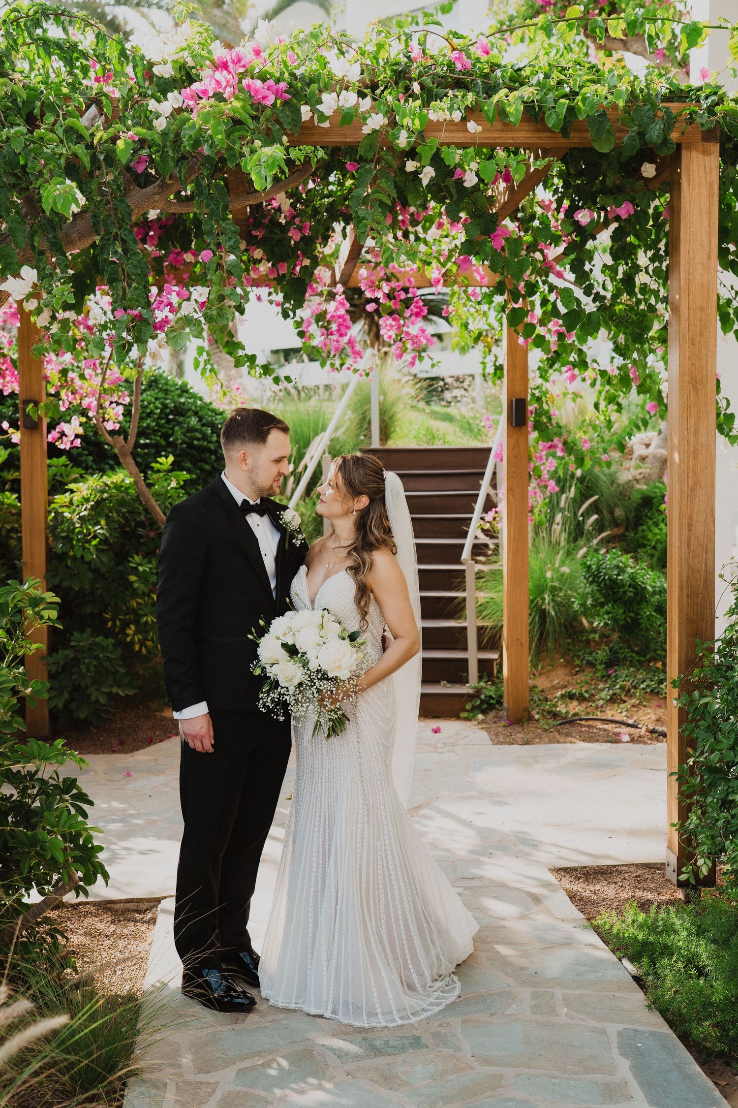 Bride and groom stand under a wooden pergola adorned with pink bougainvillea, sharing a romantic moment on their wedding day.