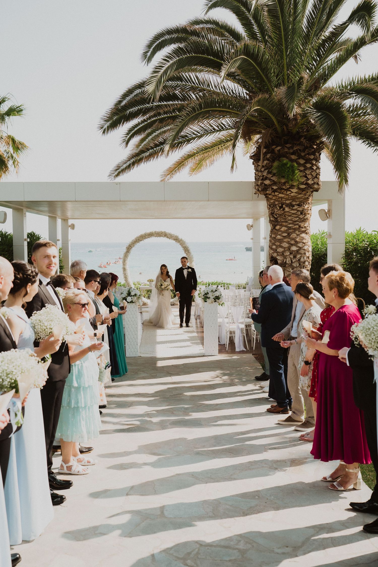 Outdoor wedding ceremony aisle with guests standing, palm trees, circular floral arch, and ocean view at a seaside venue.