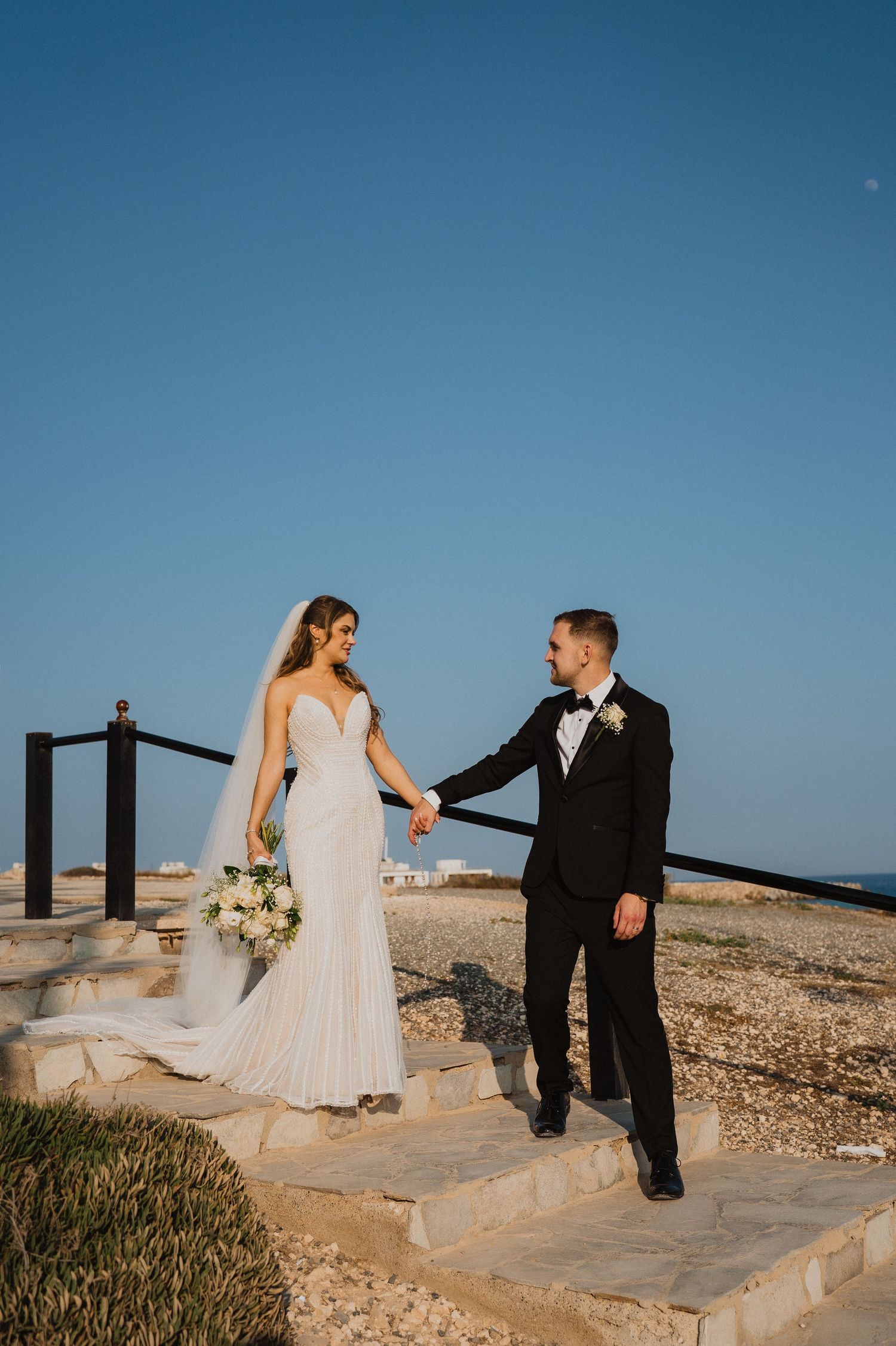 Bride and groom hold hands on stone steps by the seaside under a clear blue sky on their wedding day.