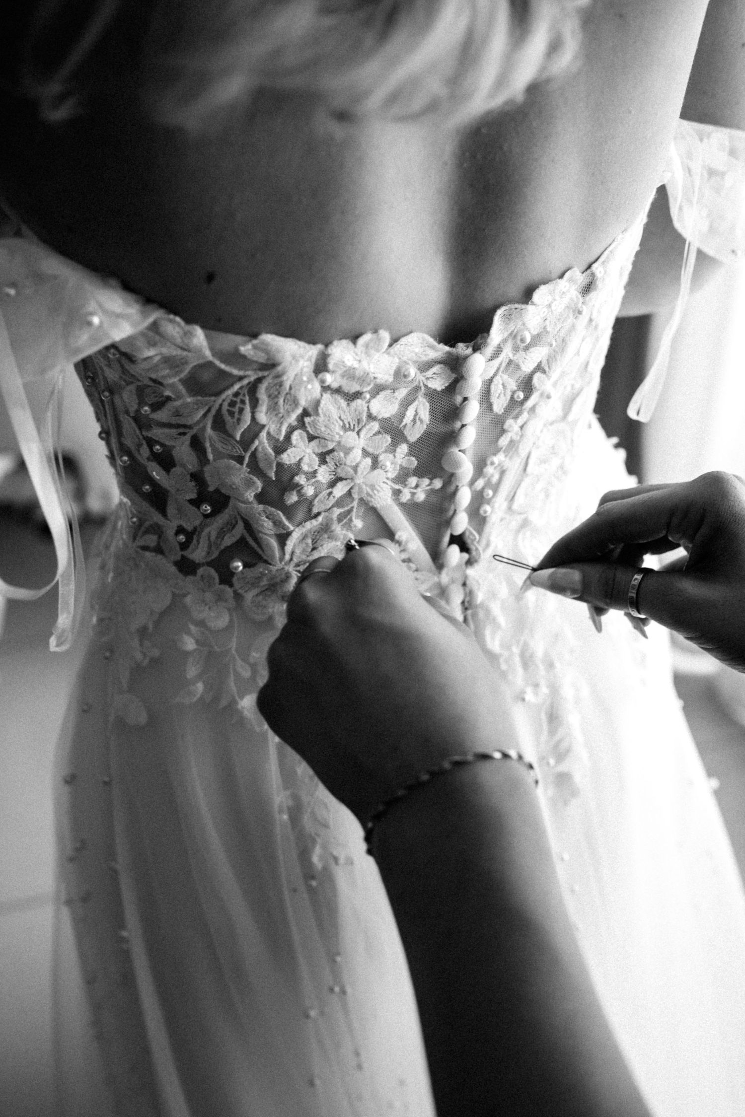 Close up black and white detail of intricate lace detailing on the back of a wedding dress being fastened.