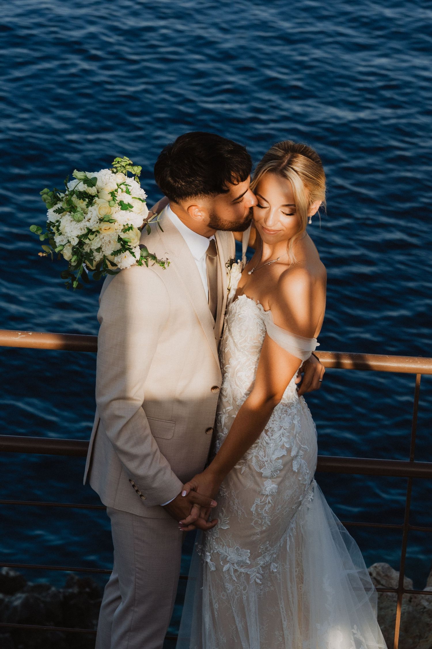 A romantic wedding photo by the ocean with a white floral bouquet and beige suit against deep blue waters.