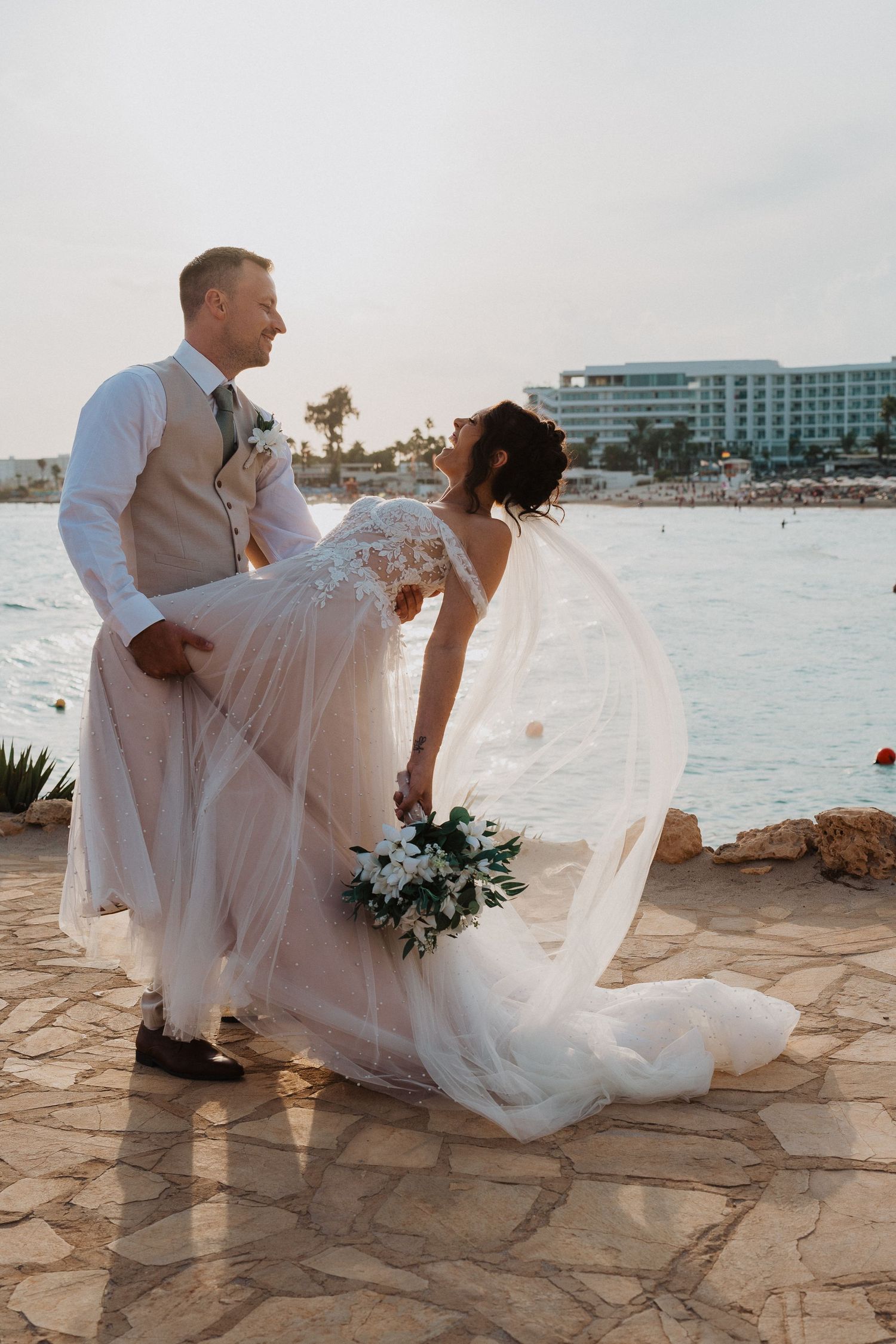 A newlywed couple poses for a romantic photo on a stone patio overlooking a beachfront resort at sunset.
