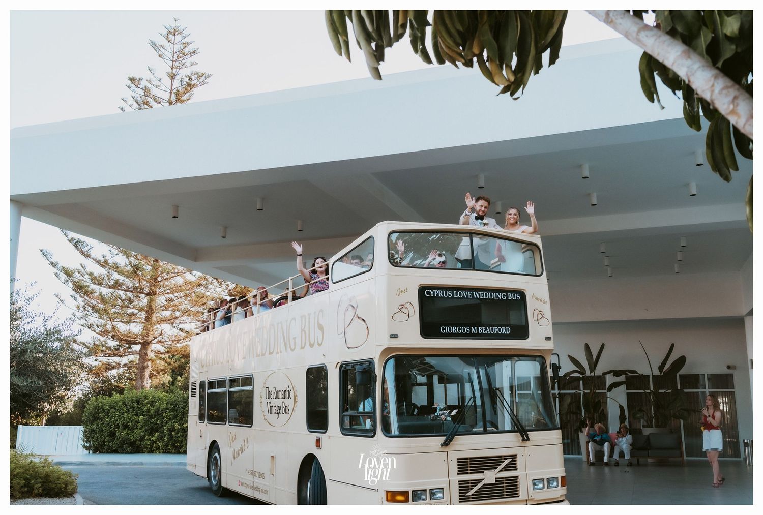 White double-decker wedding bus parked outside modern building with tropical landscaping.
