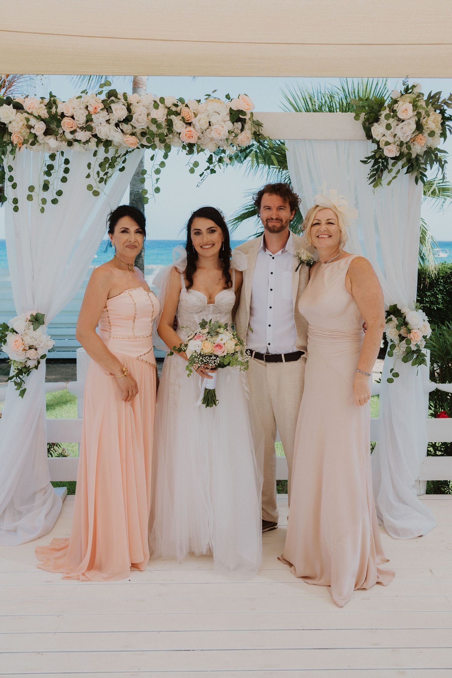 Wedding party portrait under floral arch with matching blush gowns and tropical beach backdrop.