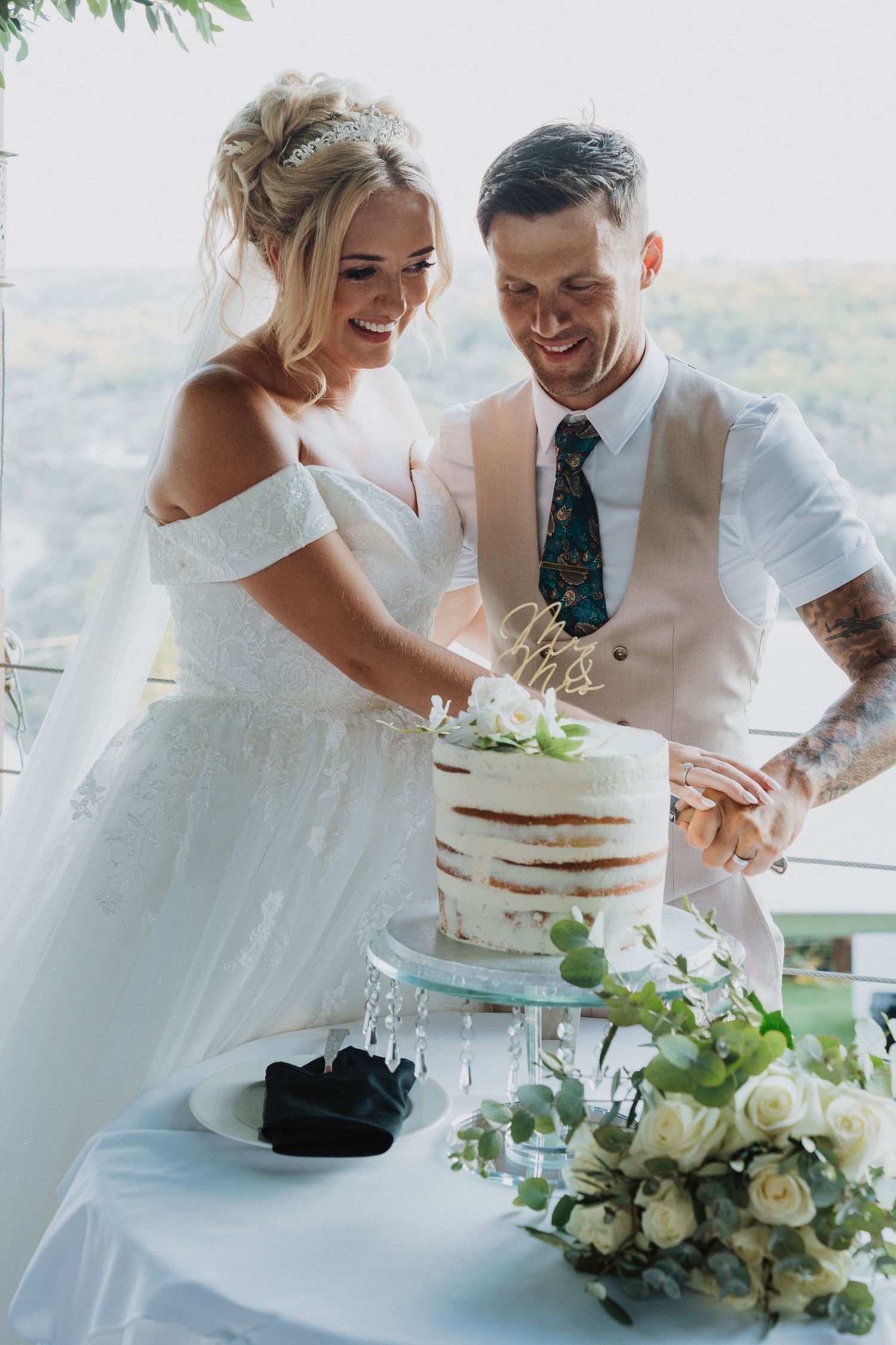 A couple cuts their elegant white naked wedding cake together at an outdoor reception venue with mountain views.