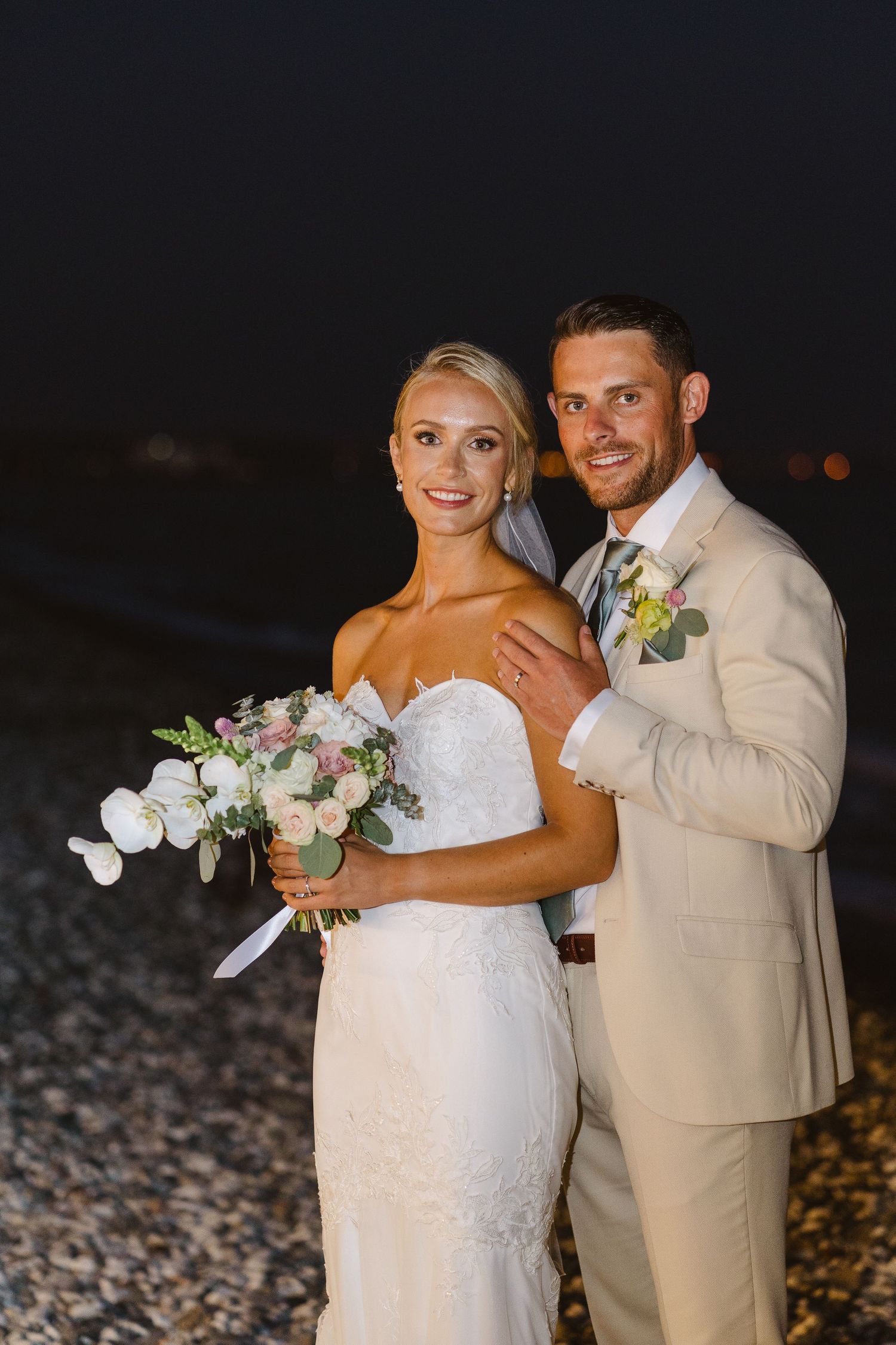 Elegant beach wedding portrait with couple in light-colored attire against dark evening sky.