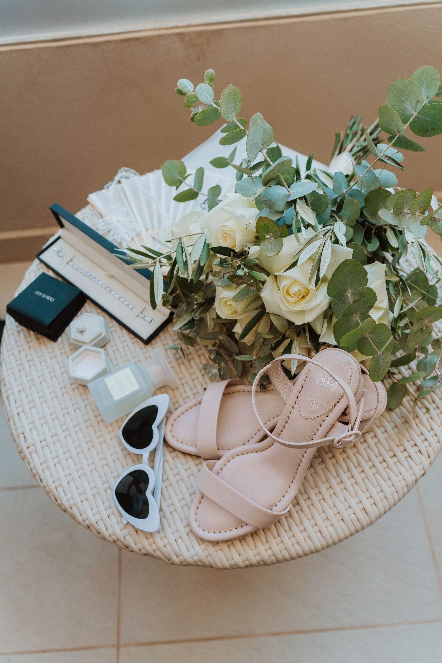 Wedding accessories including blush sandals, sunglasses, and white roses with eucalyptus arranged on a woven surface.