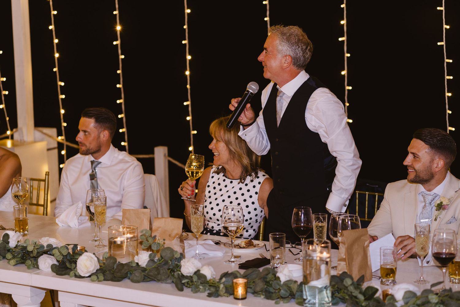 Speaker delivers toast at wedding head table decorated with floral centerpieces.