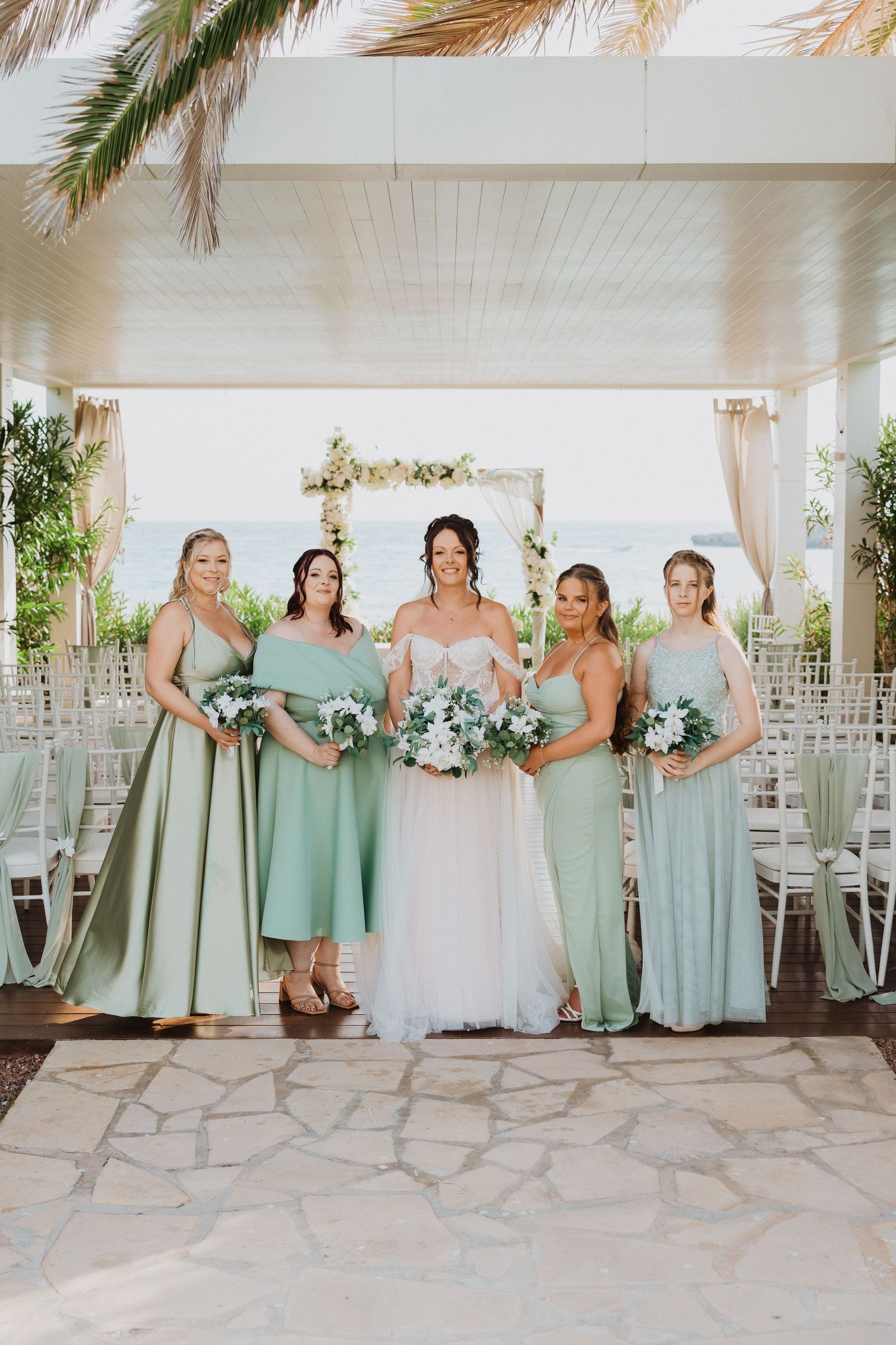Wedding party wearing elegant mint green bridesmaid dresses pose with bouquets under floral arch at outdoor venue.