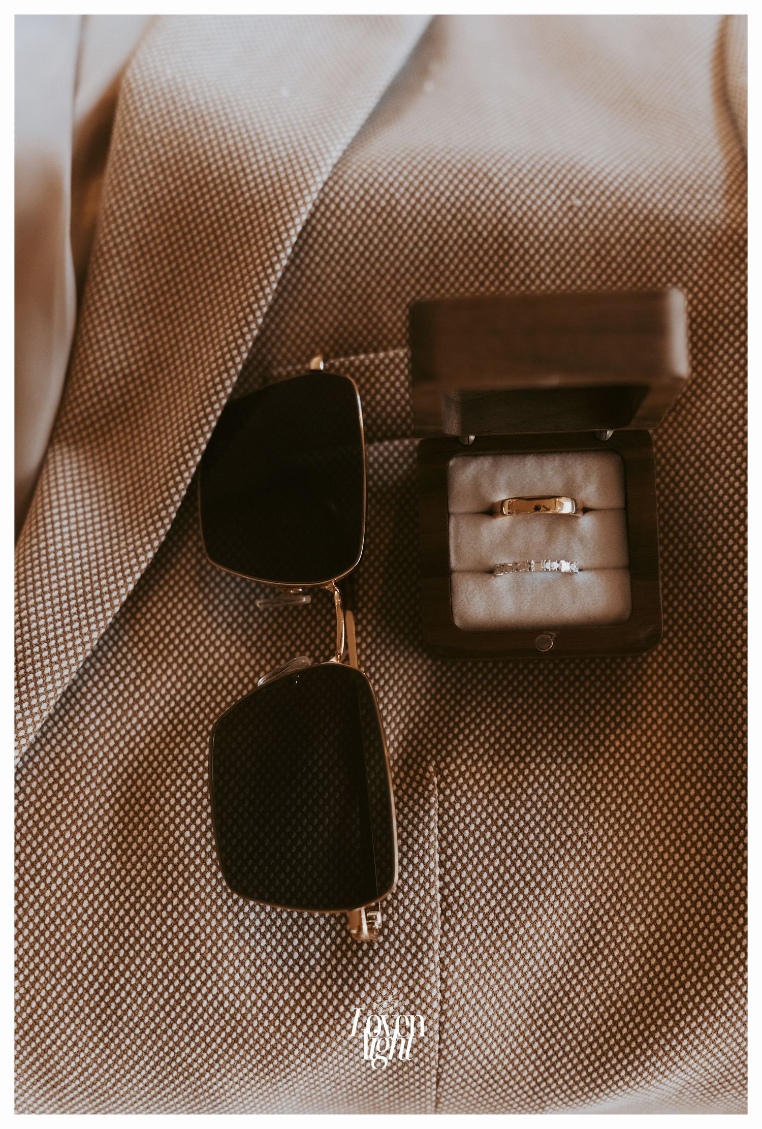 Wedding rings in a dark box alongside classic aviator sunglasses on beige fabric.