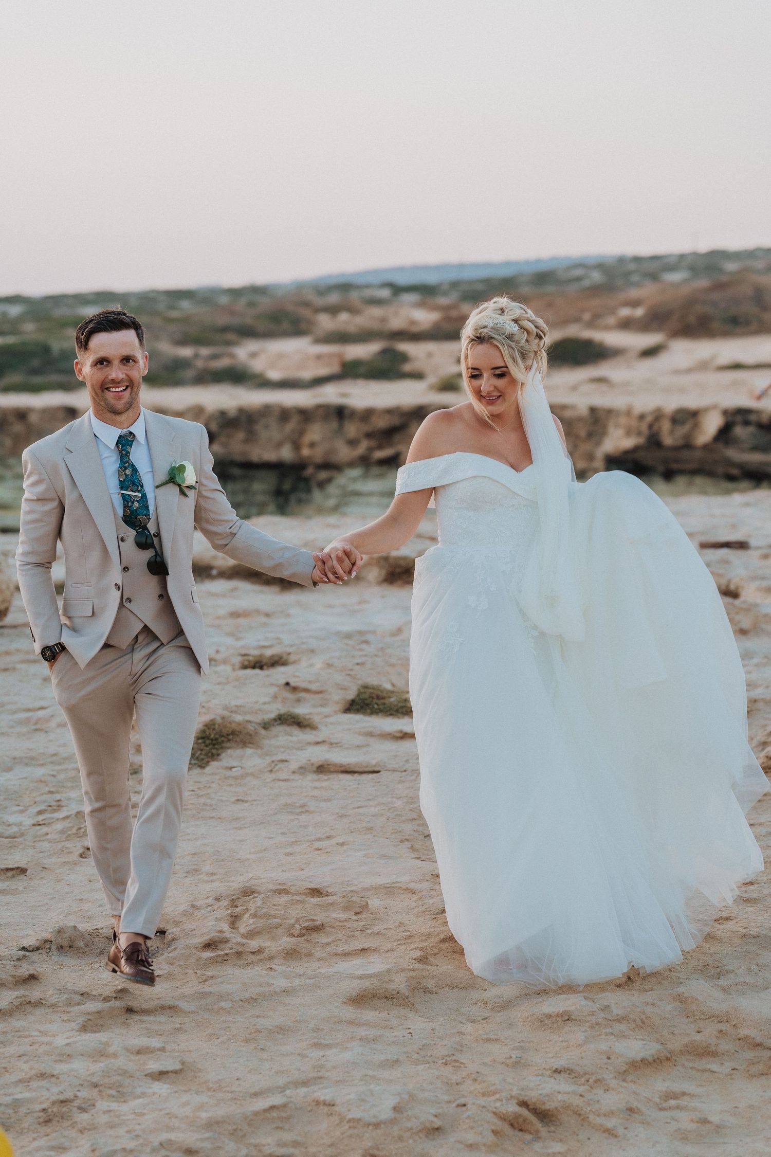 A joyful couple walks hand in hand along rocky coastal cliffs after their wedding ceremony.