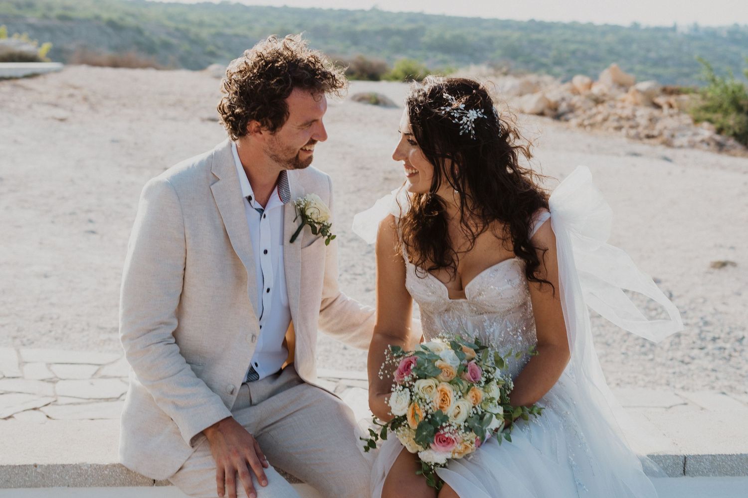A romantic wedding couple shares an intimate moment while seated on a coastal viewpoint.