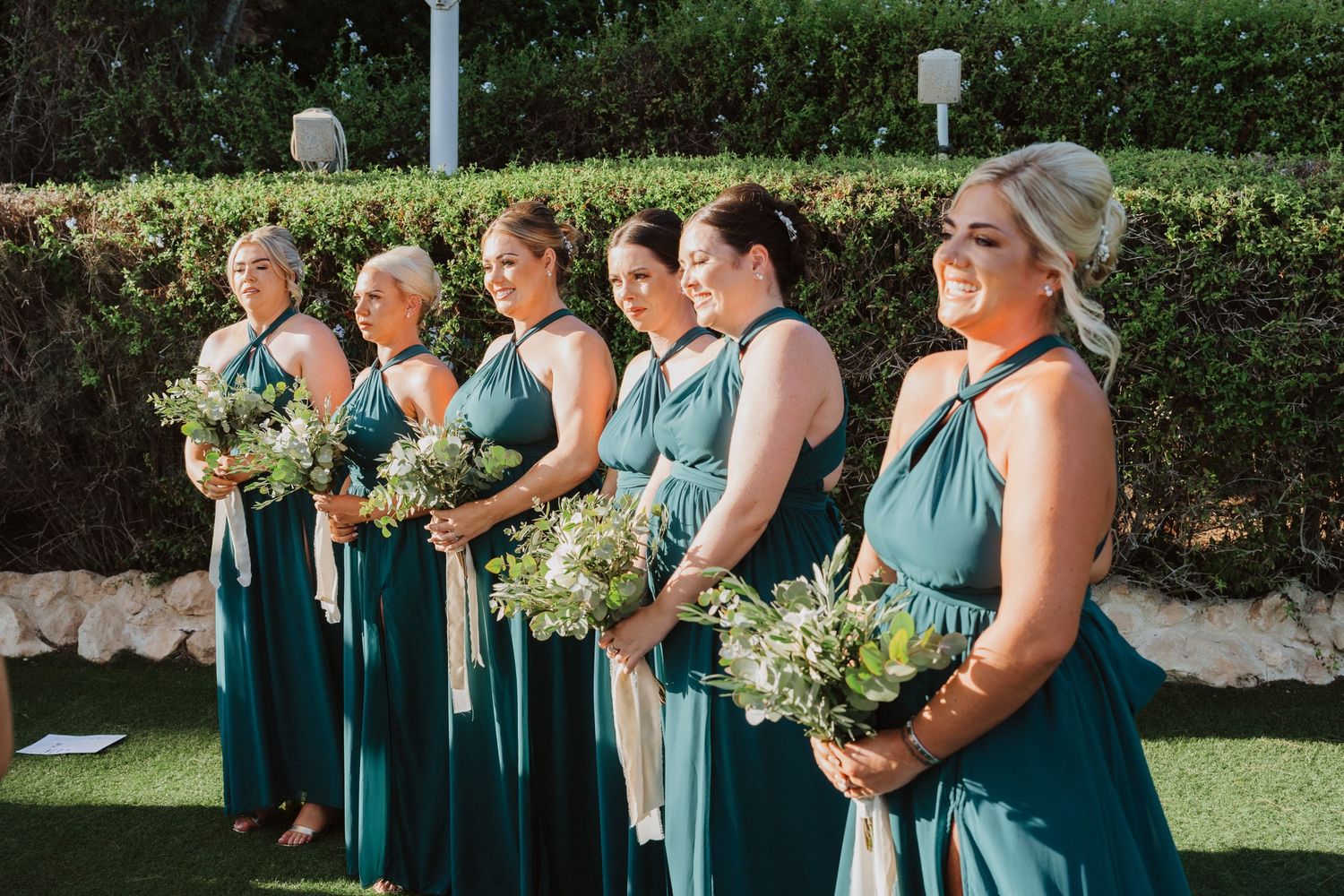 Bridesmaids in matching teal dresses stand in line holding white floral bouquets during an outdoor ceremony.