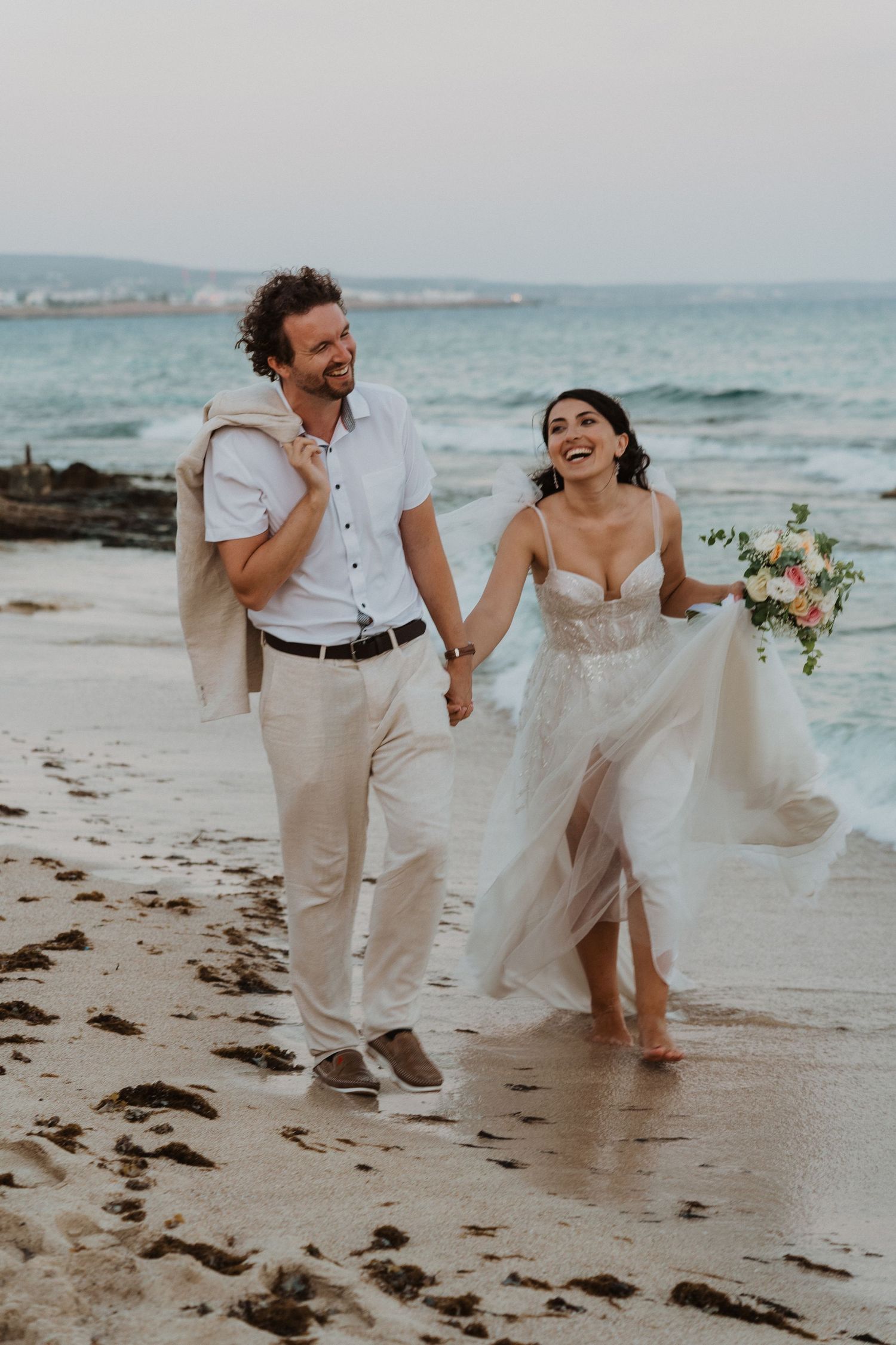 Joyful wedding couple laughing while walking barefoot on beach with waves in background.