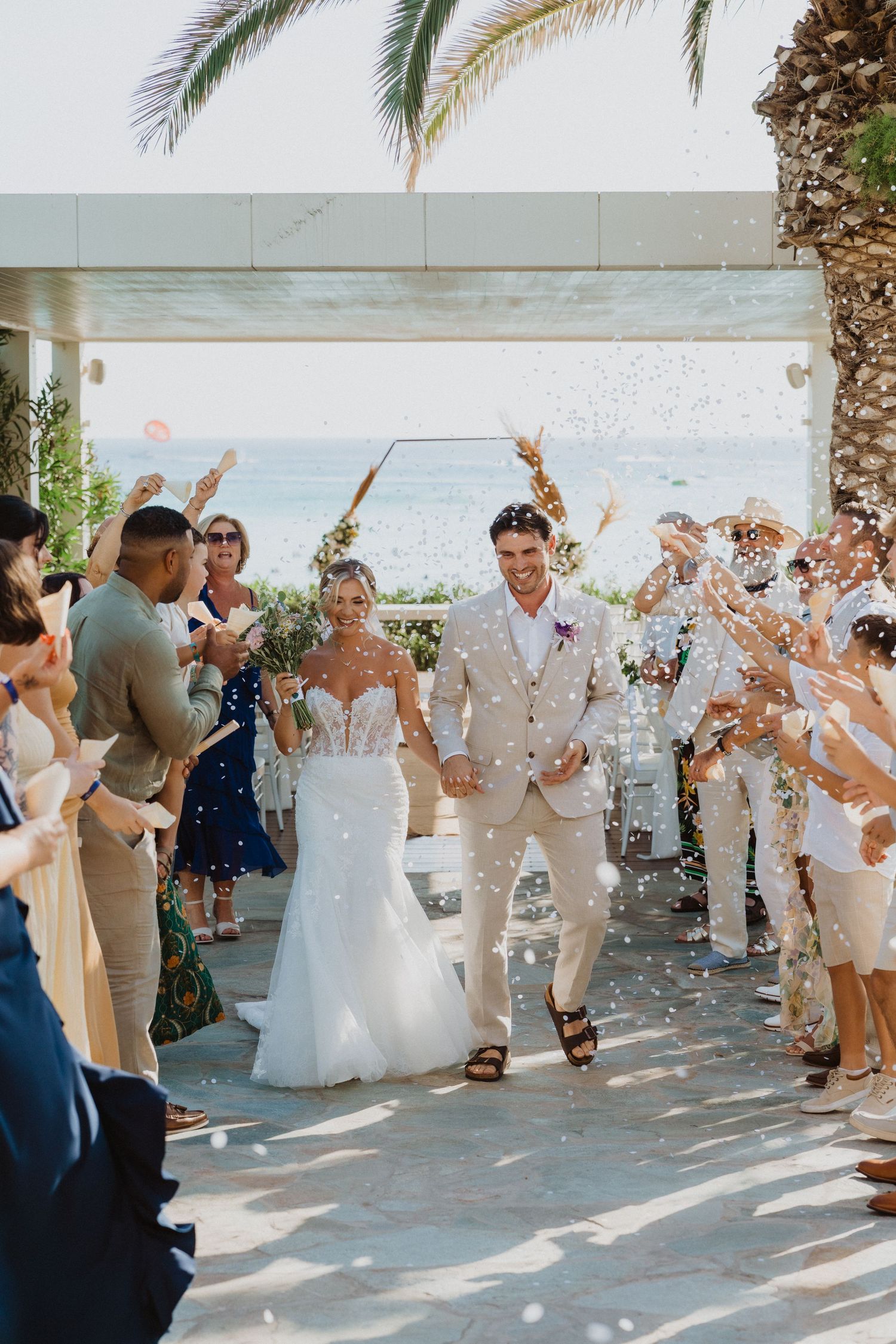 Wedding party throws confetti during celebration under palm trees.