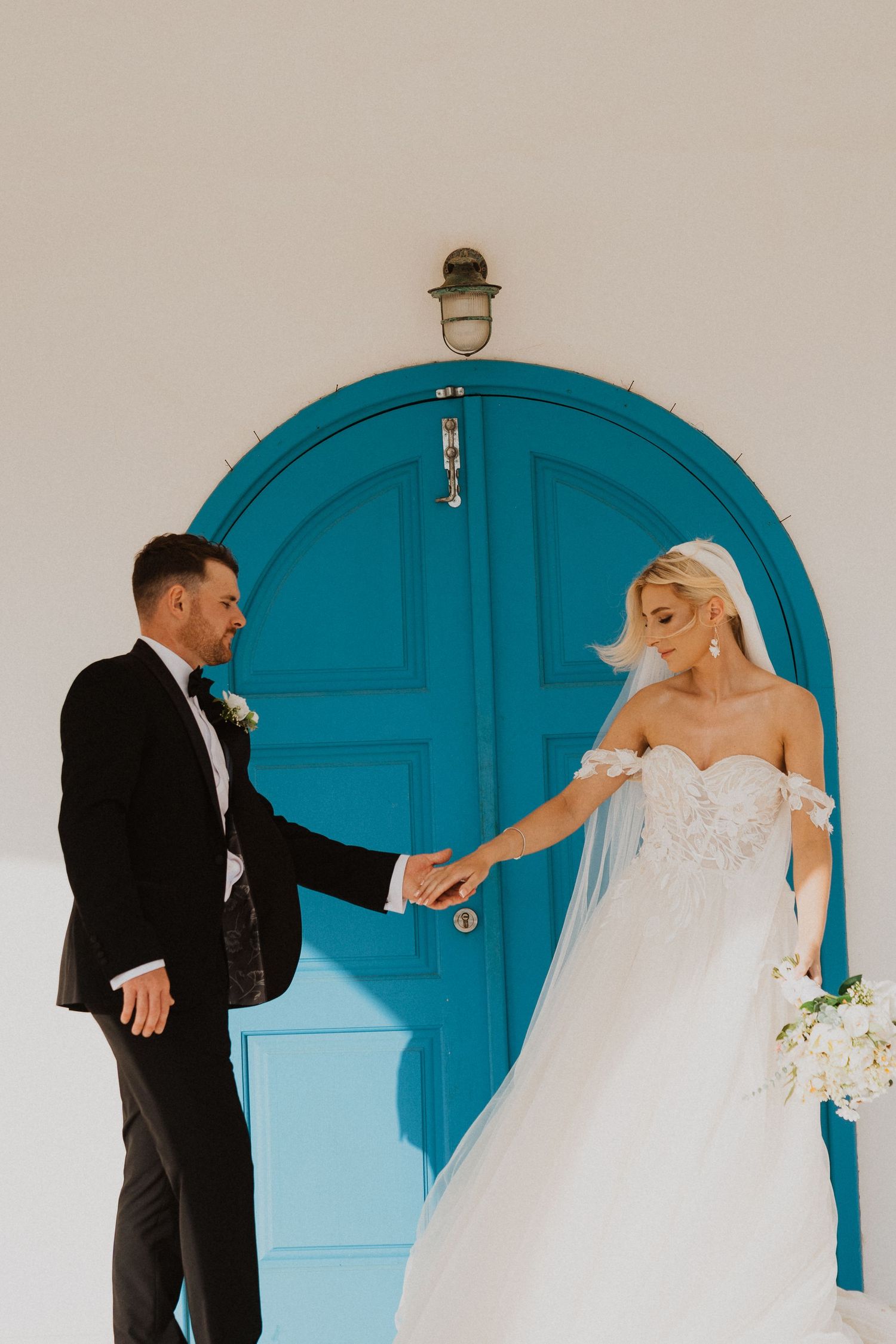 Bride and groom hold hands in front of bright blue Mediterranean church door during wedding photoshoot.