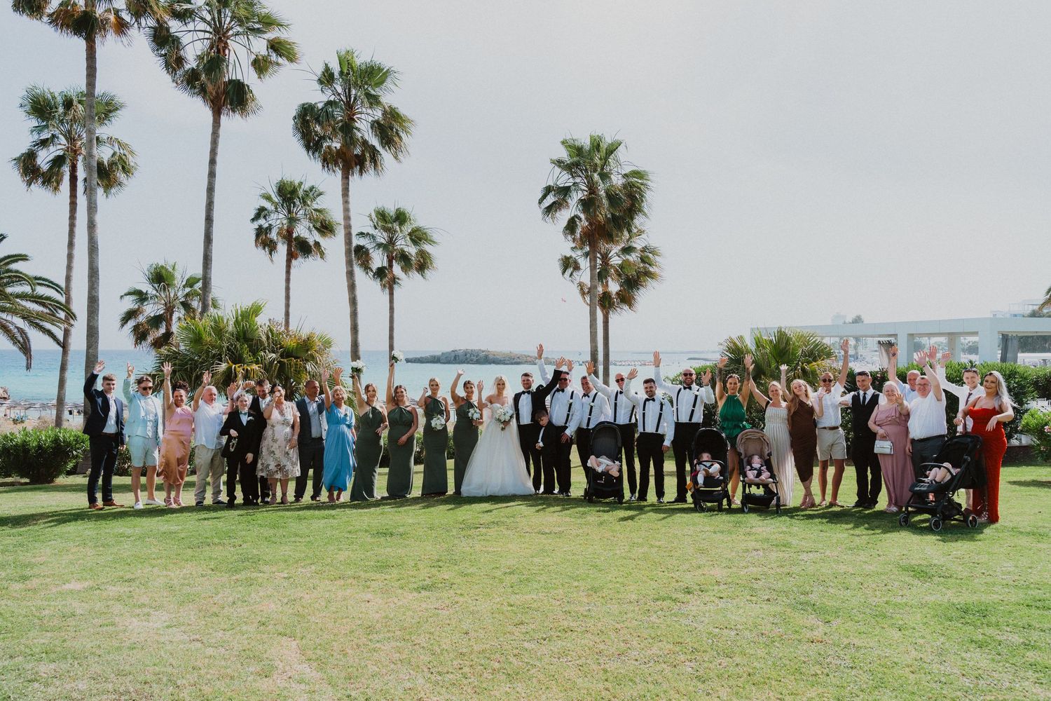 Wedding party and guests pose for group photo with palm trees and ocean in background.