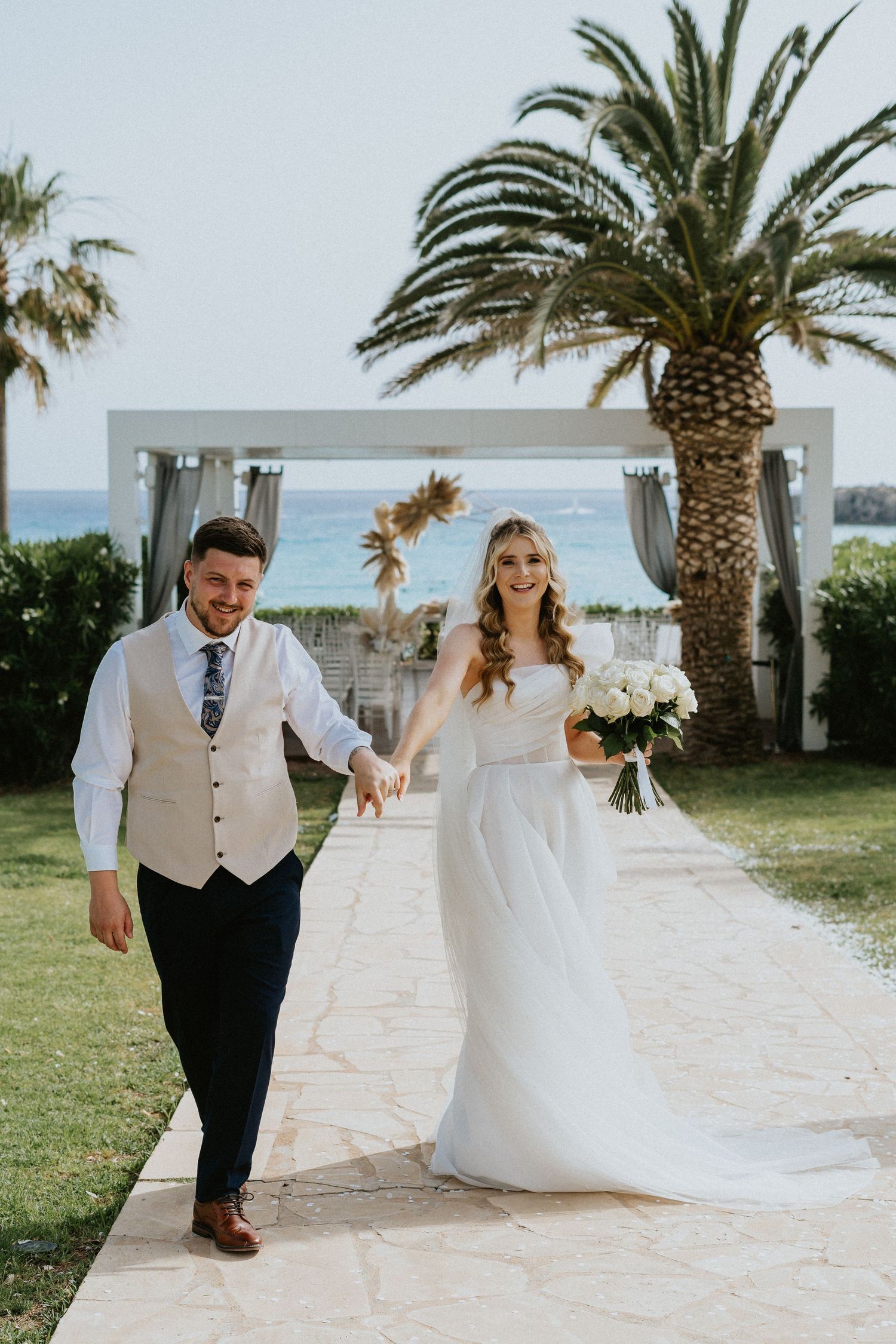 Newlyweds walk together on a beachside path with palm trees and an oceanfront wedding arch in the background.
