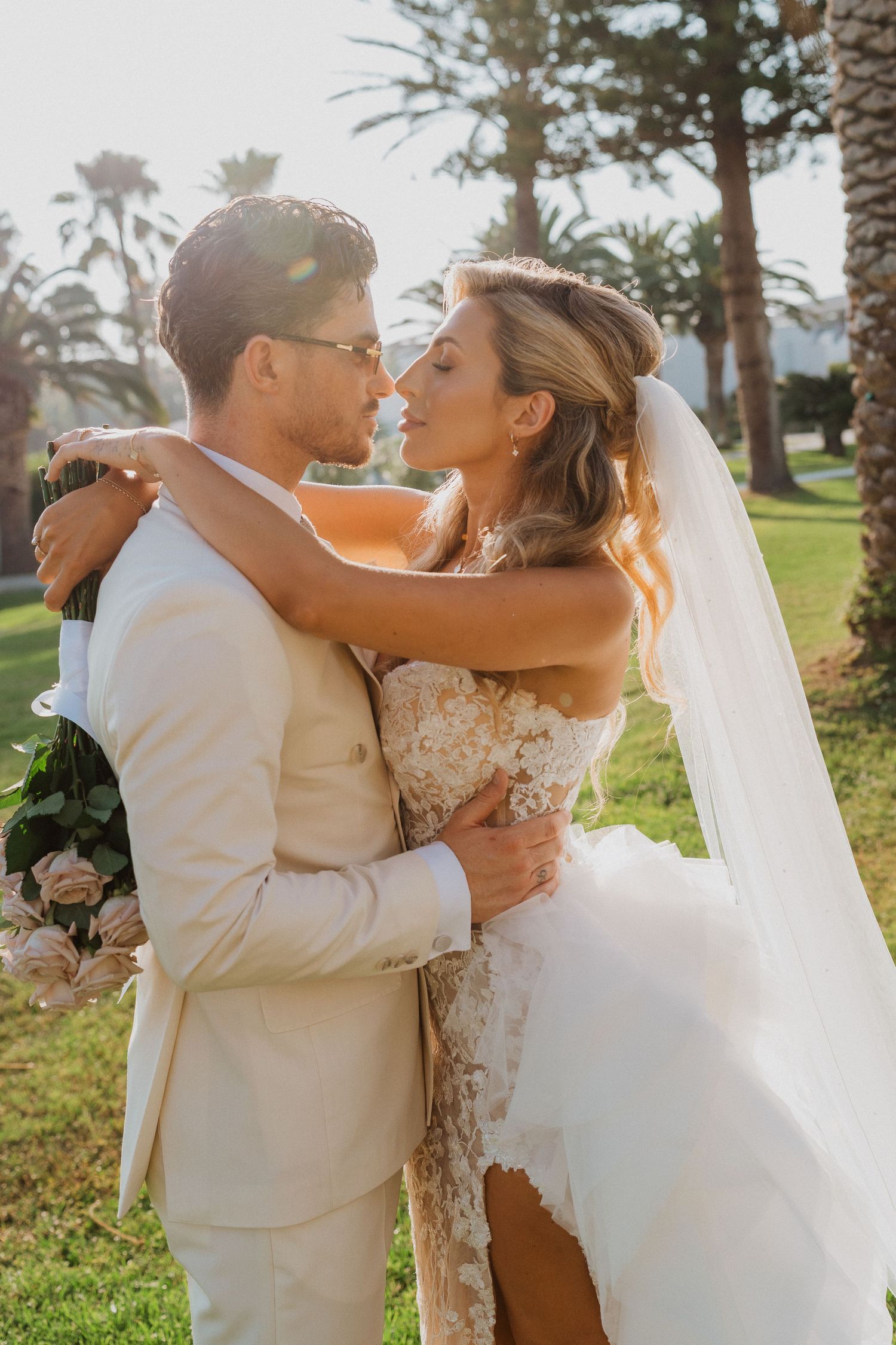 A romantic outdoor wedding scene with a couple embracing on a sunny day among palm trees and lush greenery.