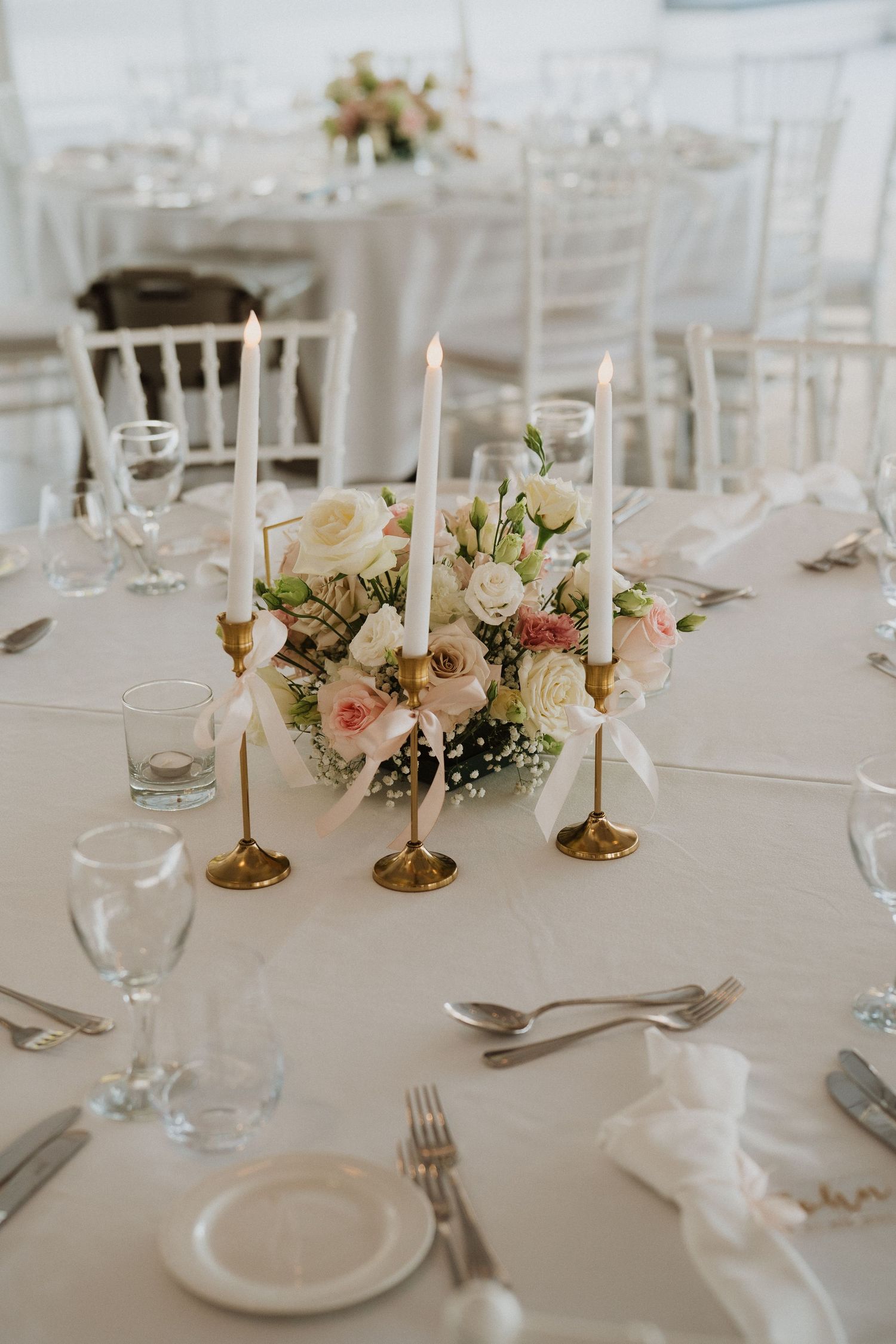 Elegant wedding reception table setting with white linens, brass candlesticks, and soft pink floral centerpieces.