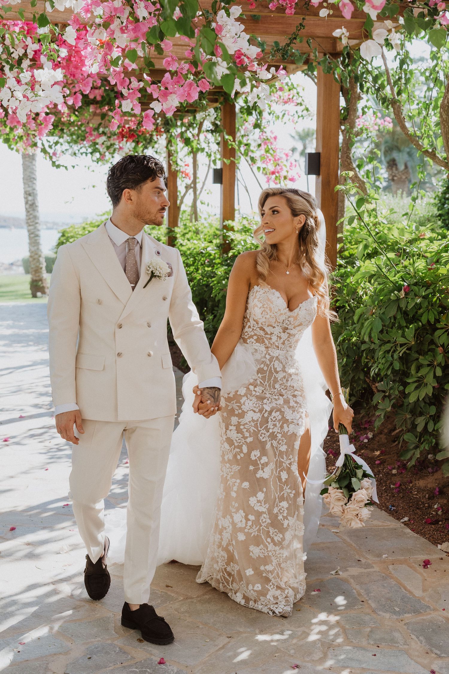 A couple in wedding attire walks under a pink floral archway in an outdoor garden setting.