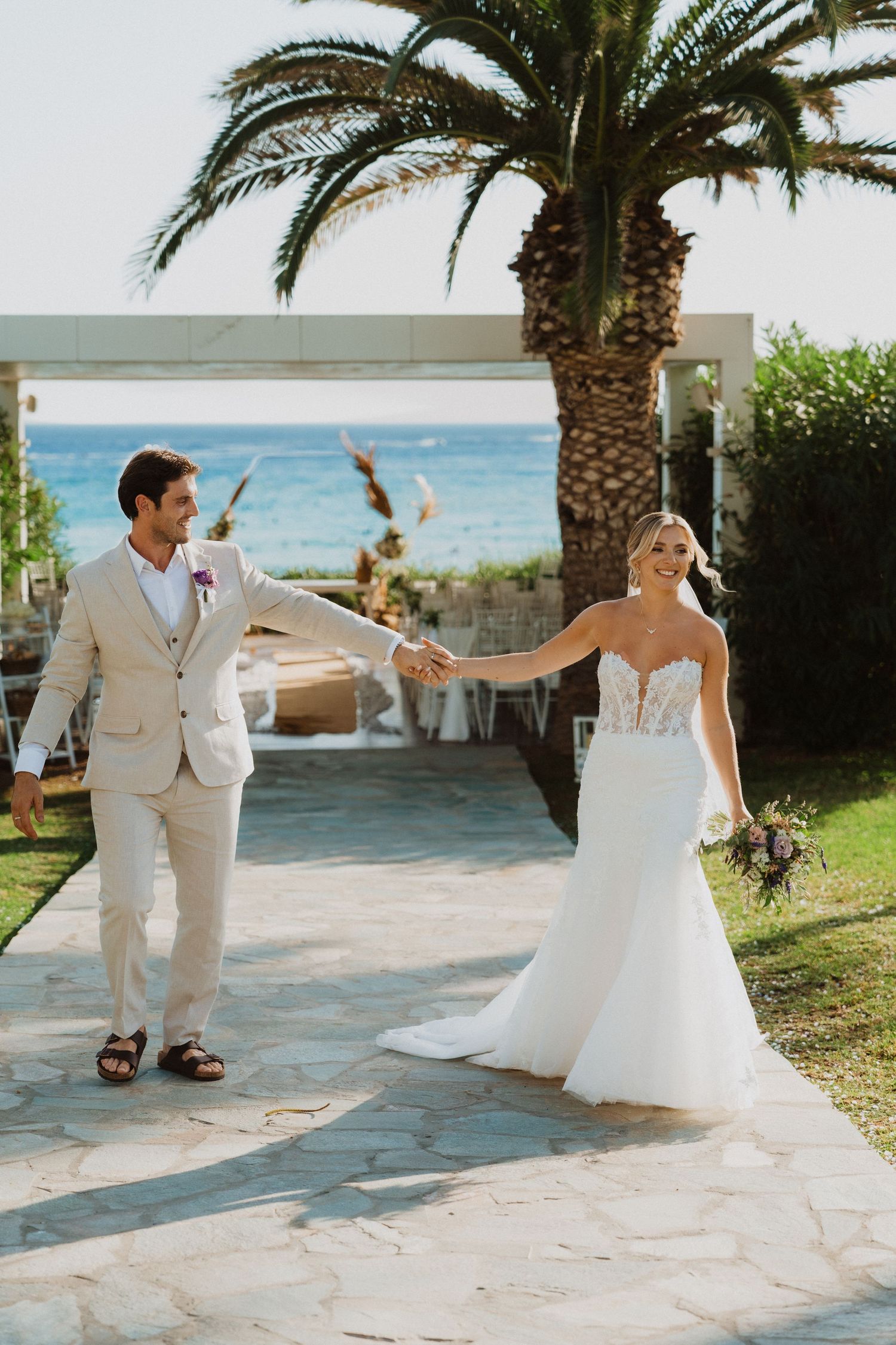 A couple in wedding attire dances joyfully on a stone path with a palm tree and ocean view in the background.