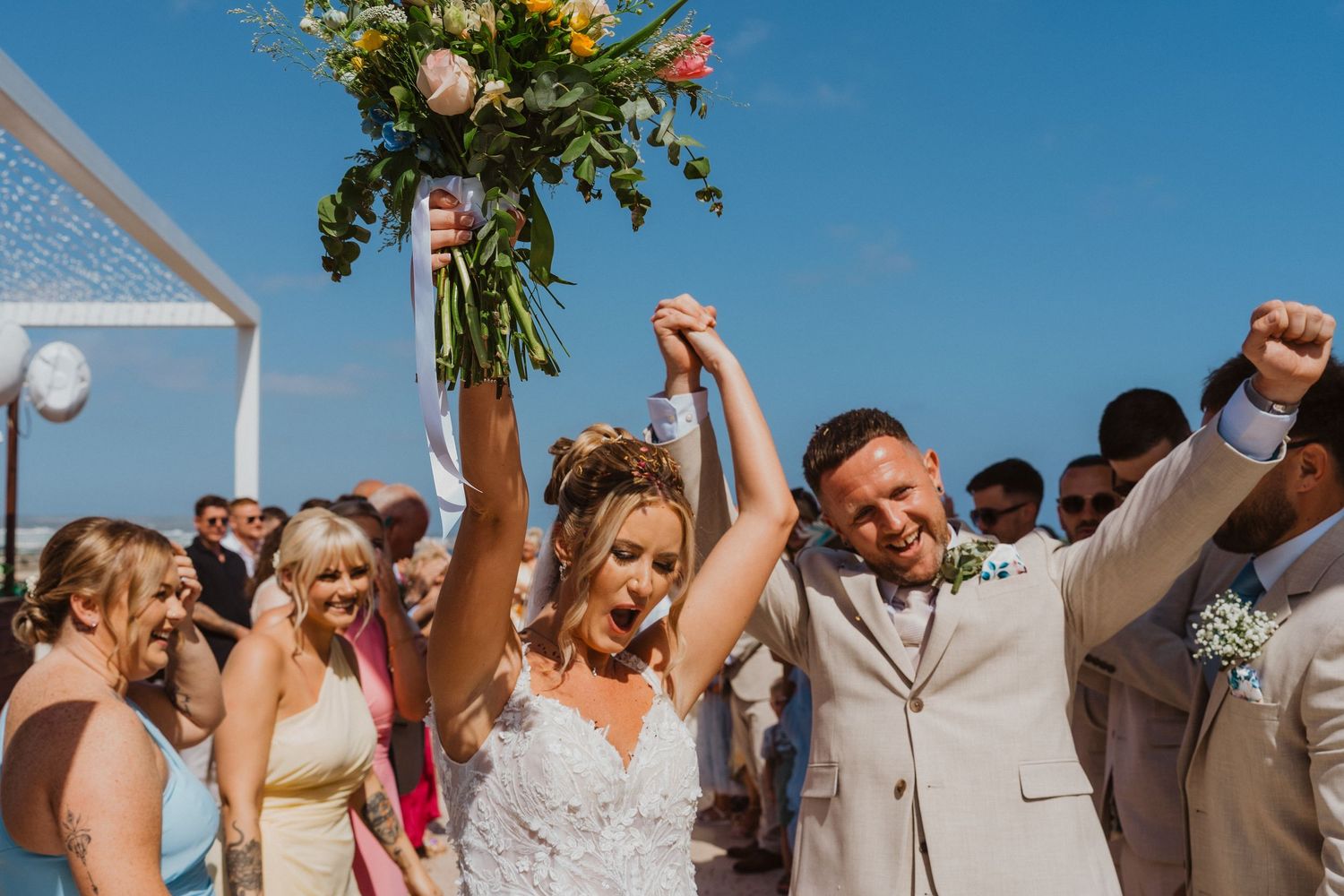 Wedding guests cheer and raise their arms in celebration during an outdoor ceremony against a bright blue sky.