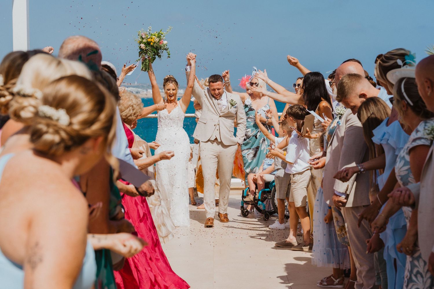 Wedding guests dance in a circle on a sunny beach during a seaside celebration.