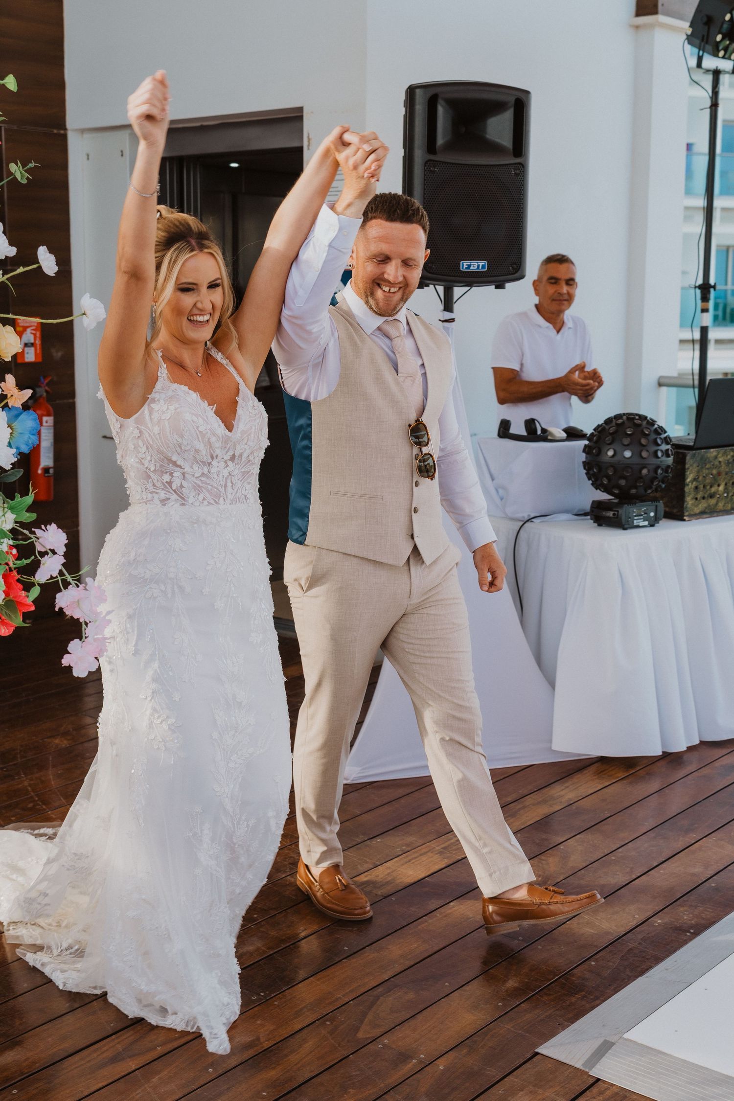 A couple celebrates joyfully on their wedding day, dancing together on a wooden deck with a DJ booth visible in background.
